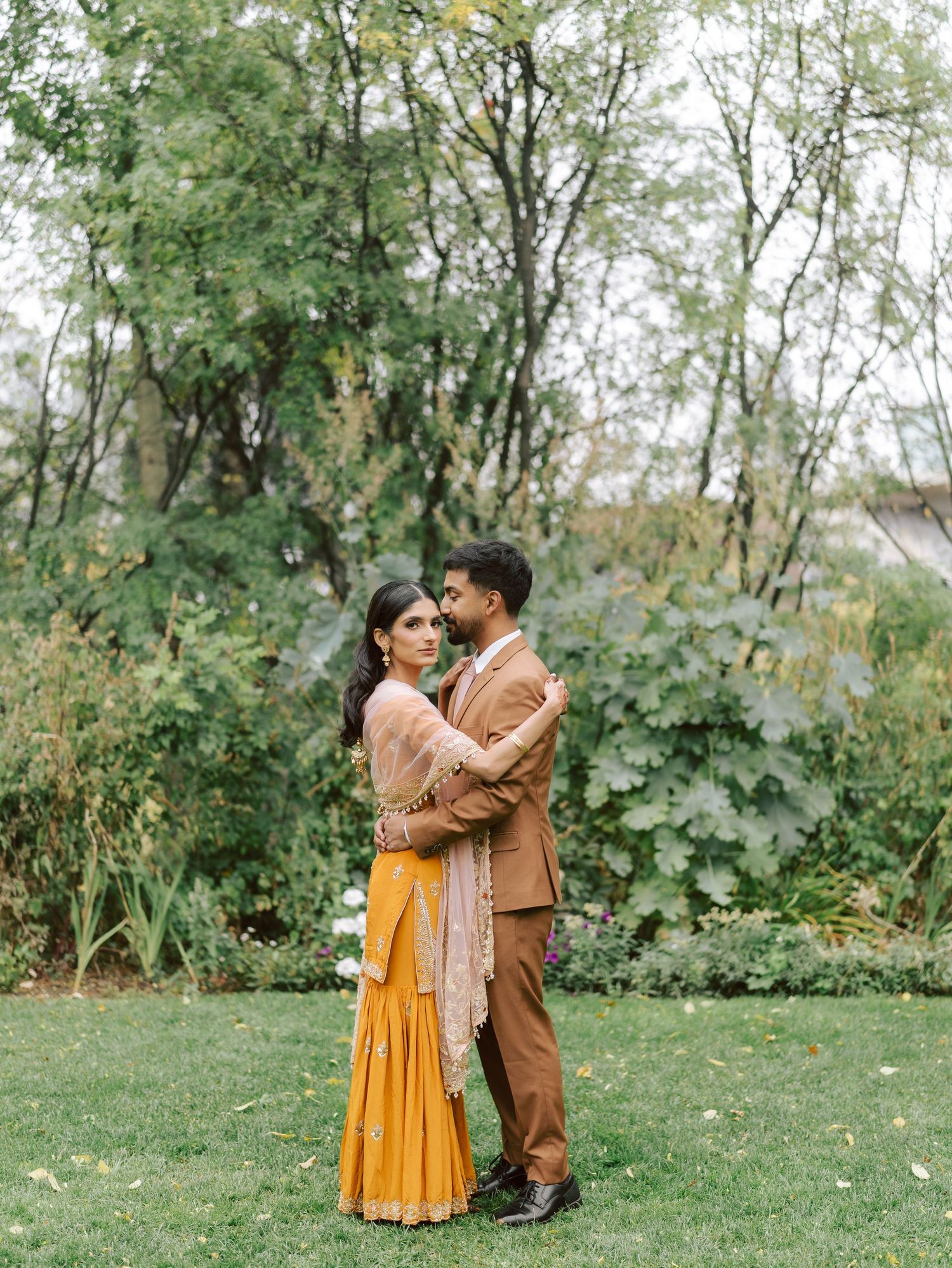 South Asian bride and groom embrace romantically in lush garden during traditional wedding photoshoot.