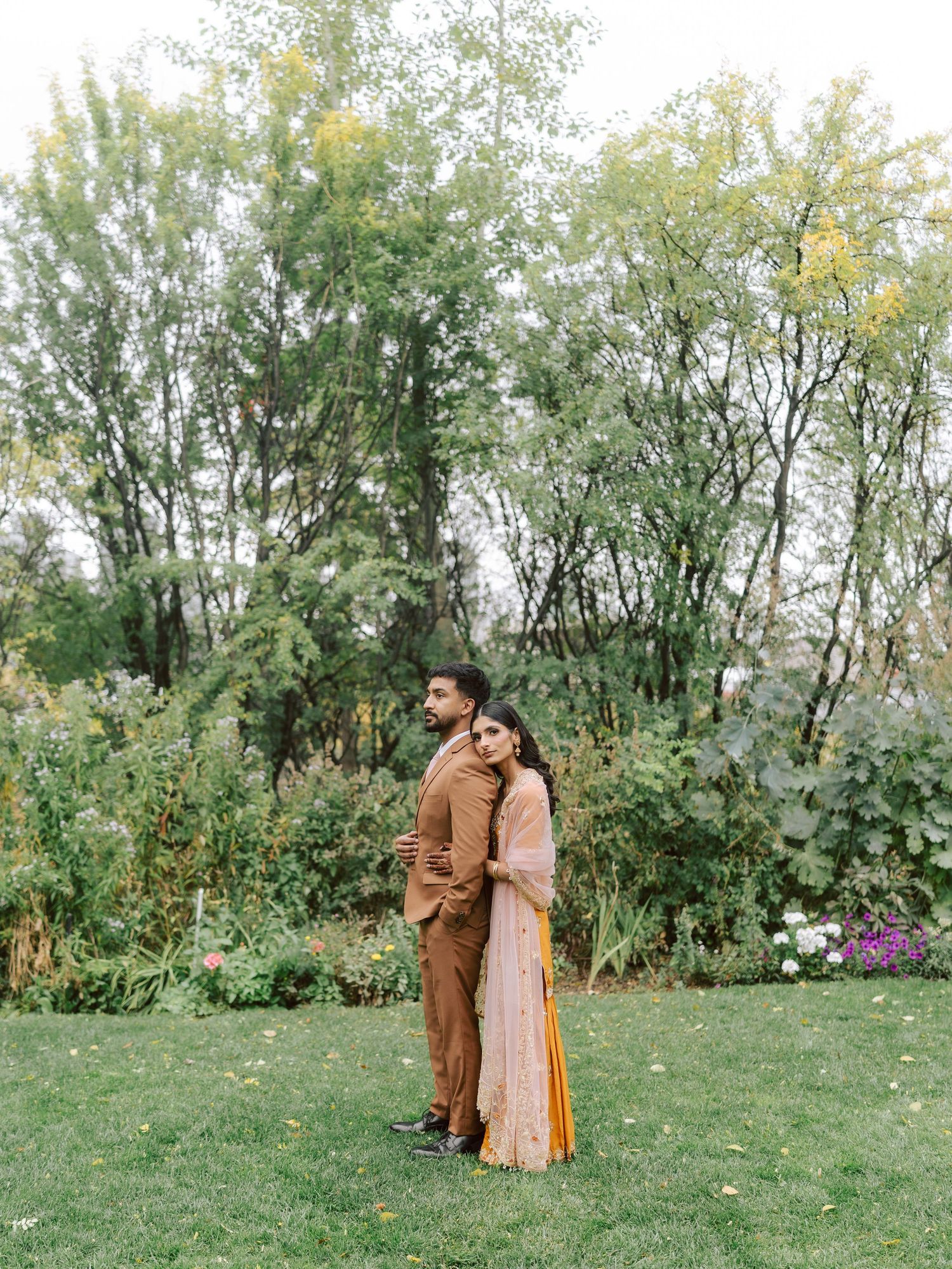 South Asian couple embracing in autumn garden, bride in pink dupatta over yellow lehenga with groom.