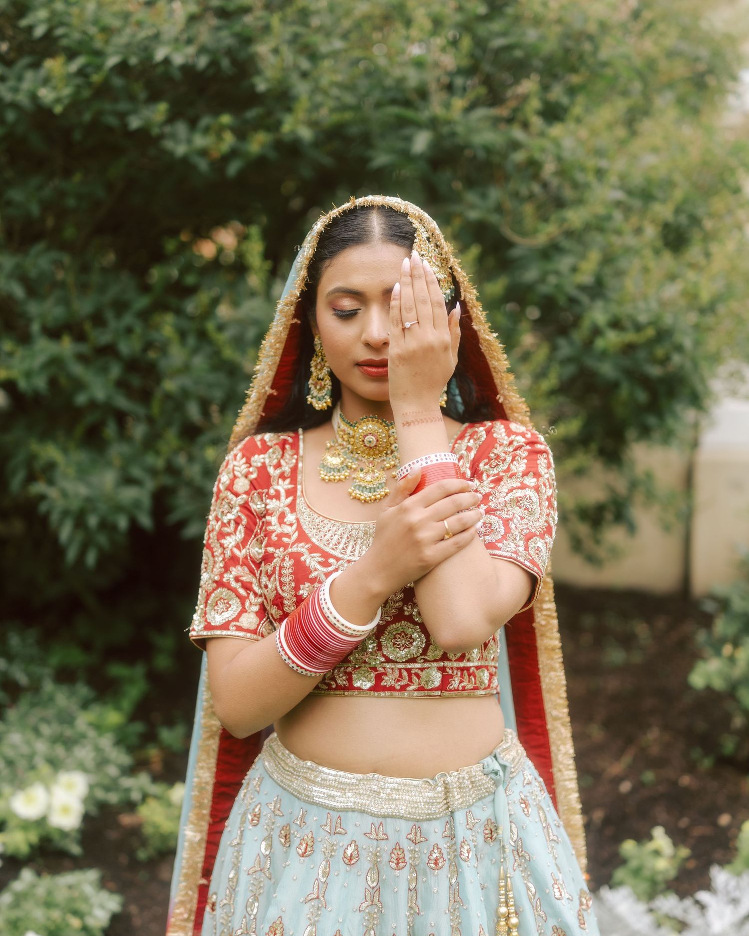 Indian bride in red and blue lehenga with gold jewelry poses gracefully outdoors.