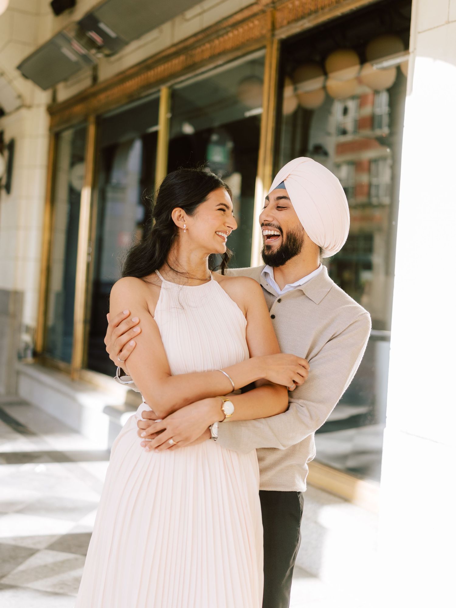 Laughing Sikh couple embracing outside elegant storefront, woman in white halter dress.