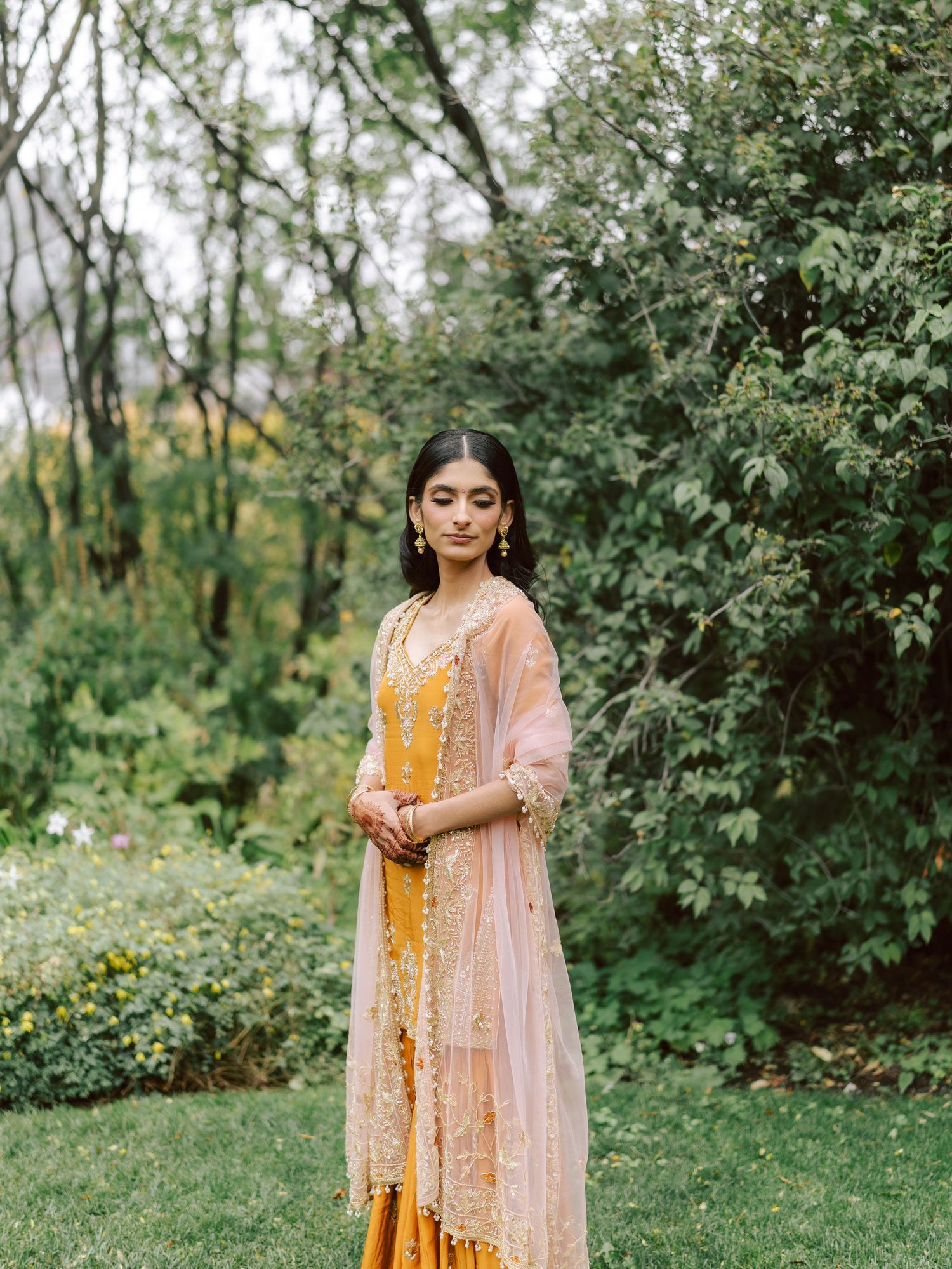 South Asian bride in yellow sharara with eyes closed stands serenely in lush green garden.