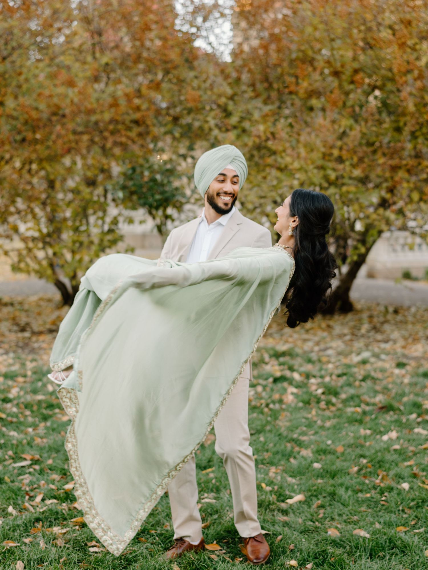 Sikh groom in mint turban joyfully lifting bride wrapped in flowing green dupatta among fall trees.
