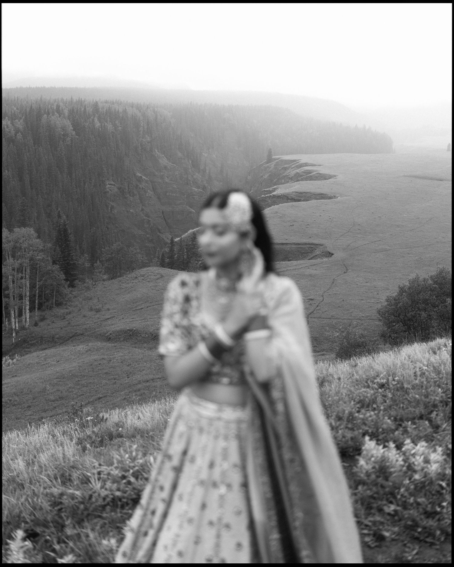 Black and white photo of Sikh bride in lehenga standing on hilltop with dramatic valley landscape.