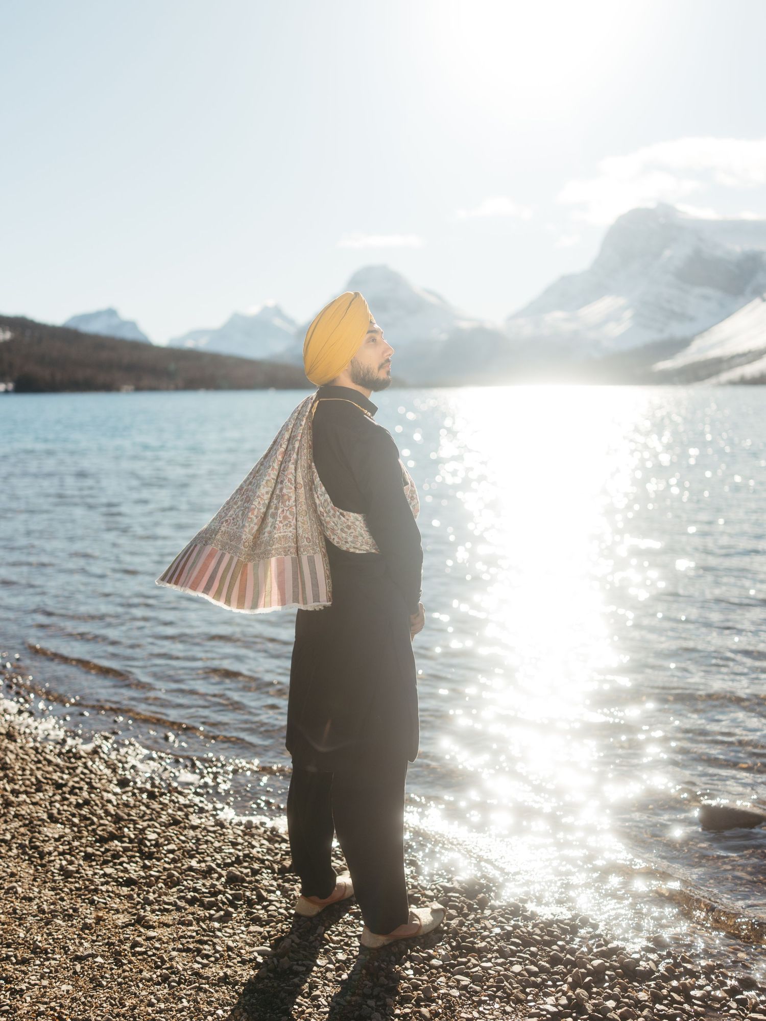 Sikh man in yellow turban and black kurta standing by mountain lake in bright sunlight.