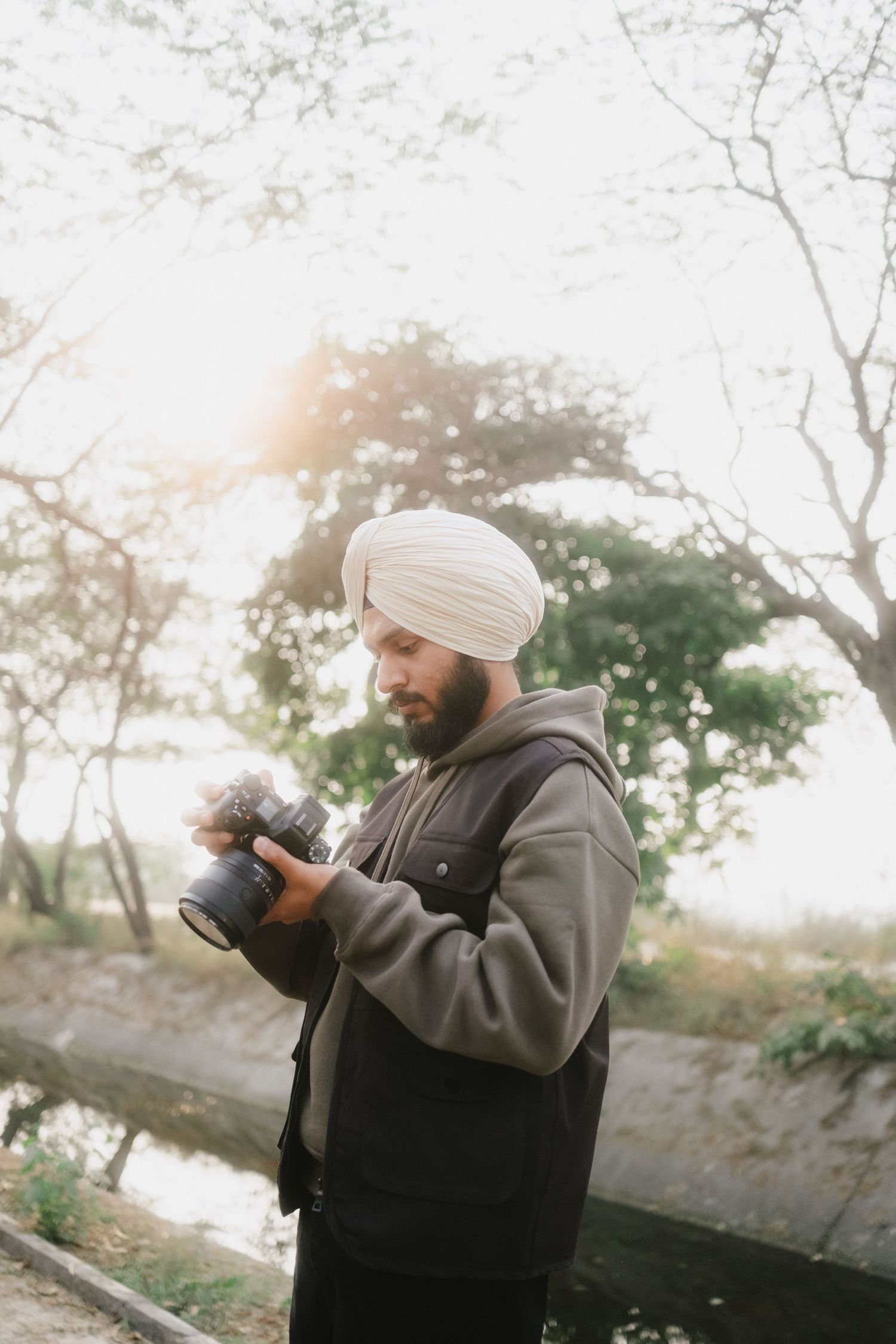 Sikh photographer in white turban reviews camera screen in sunlit outdoor forest setting.