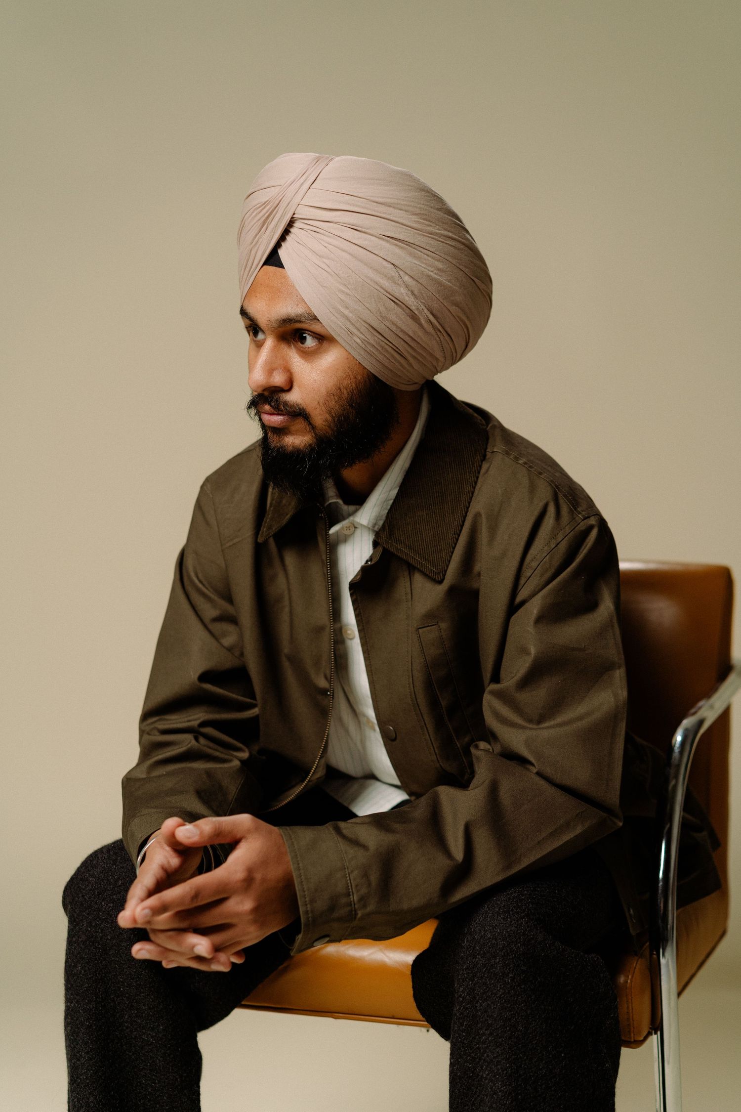 Sikh man in beige turban and olive jacket seated in leather chair against neutral background.