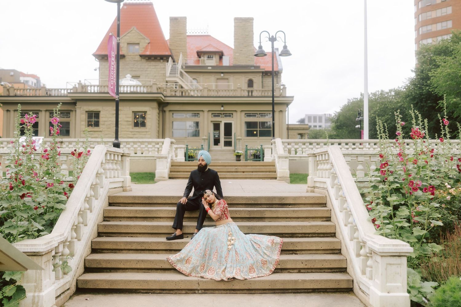 Sikh wedding couple poses on ornate staircase outside historic mansion.