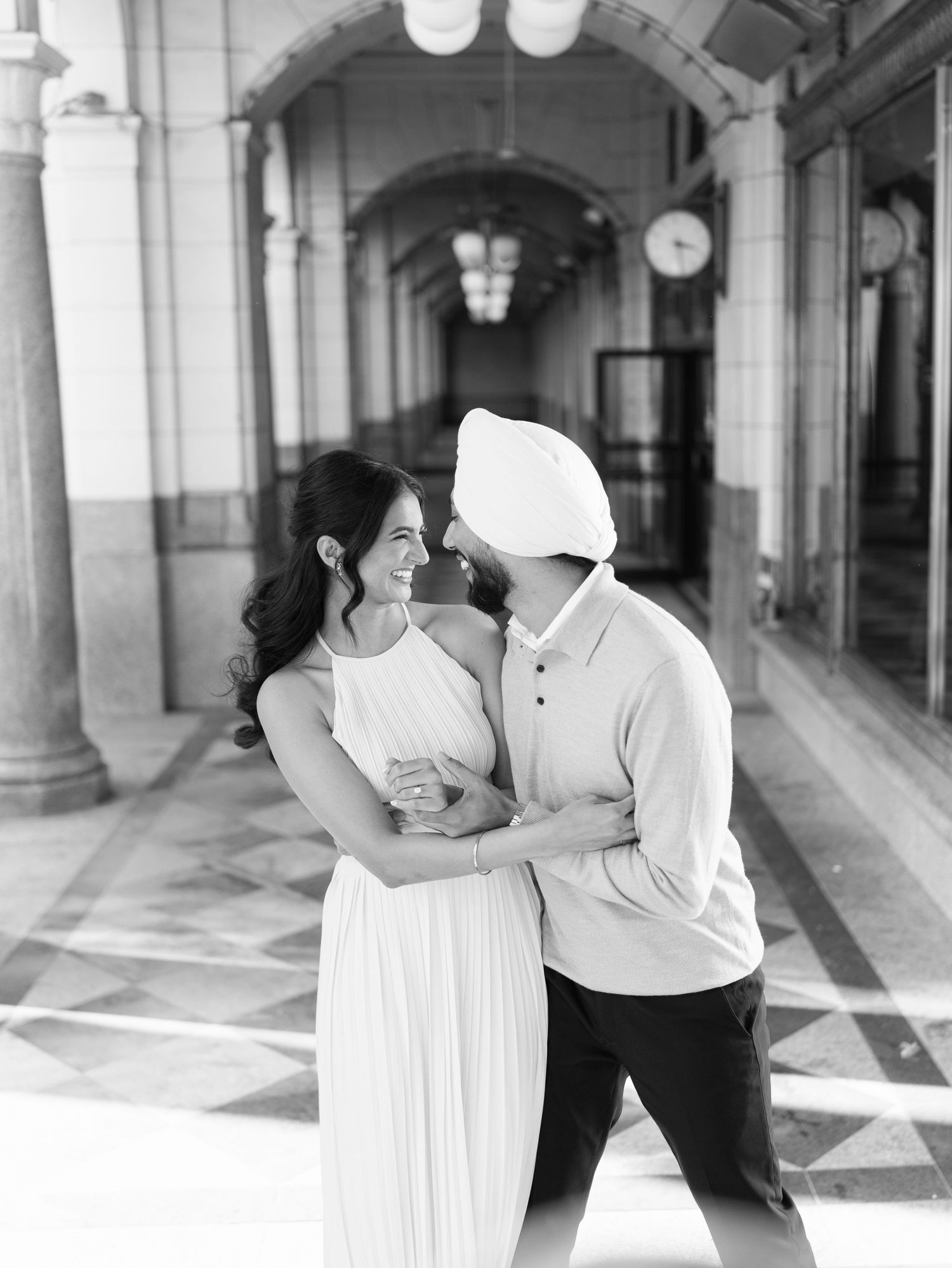 Black and white photo of smiling couple holding each other in a grand columned hallway.