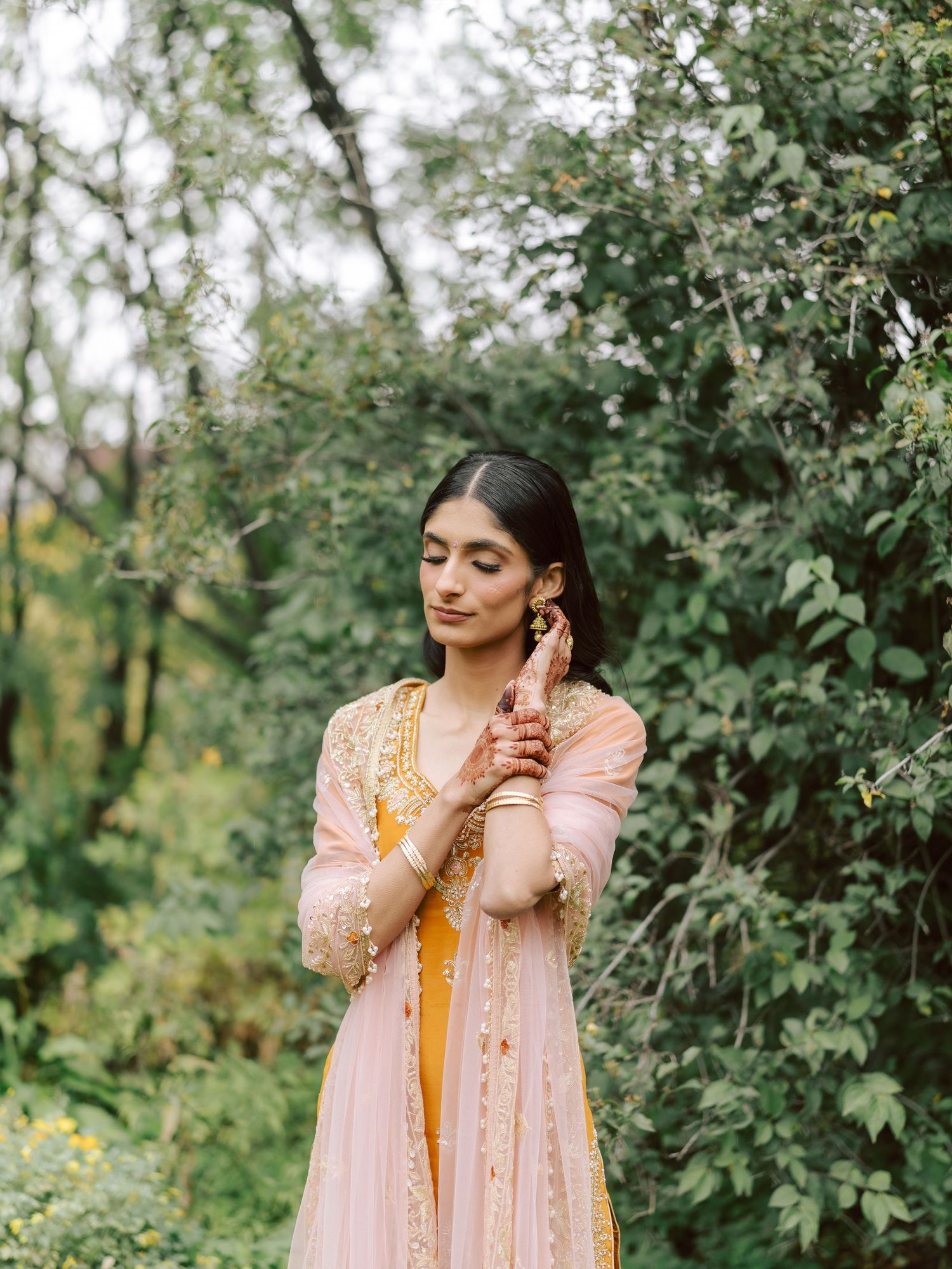 South Asian bride in pink and yellow lehenga with gold jewelry poses in lush green garden.