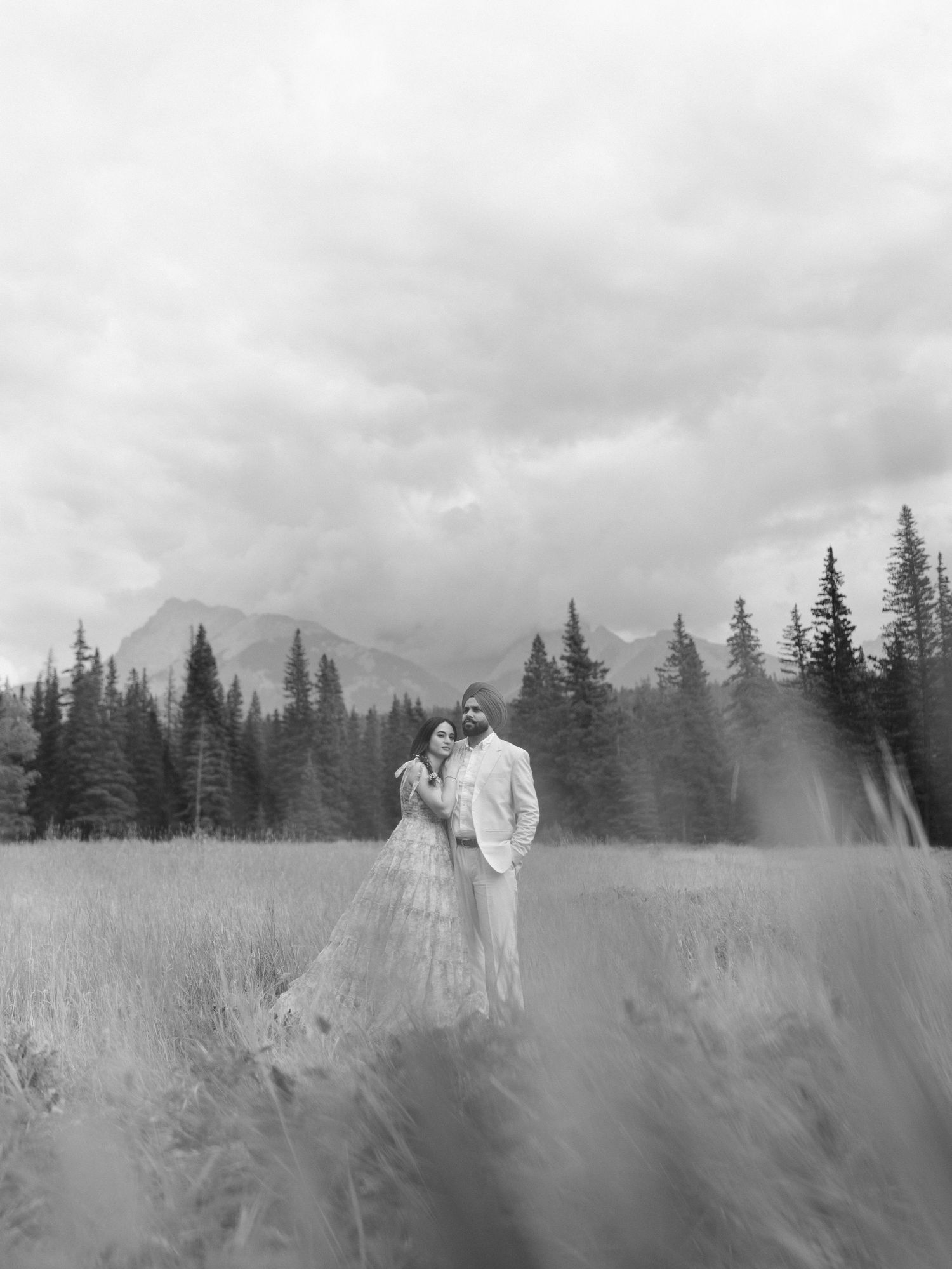 Black and white photo of a couple embracing in a mountain meadow with evergreen trees and dramatic cloudy sky.