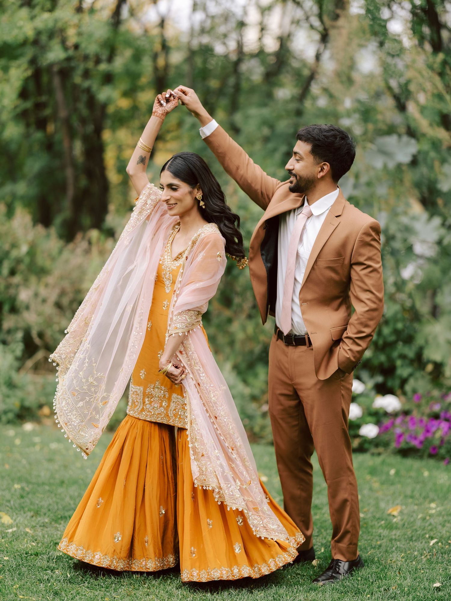 Groom twirls bride in yellow sharara and blush dupatta during outdoor South Asian wedding photoshoot.
