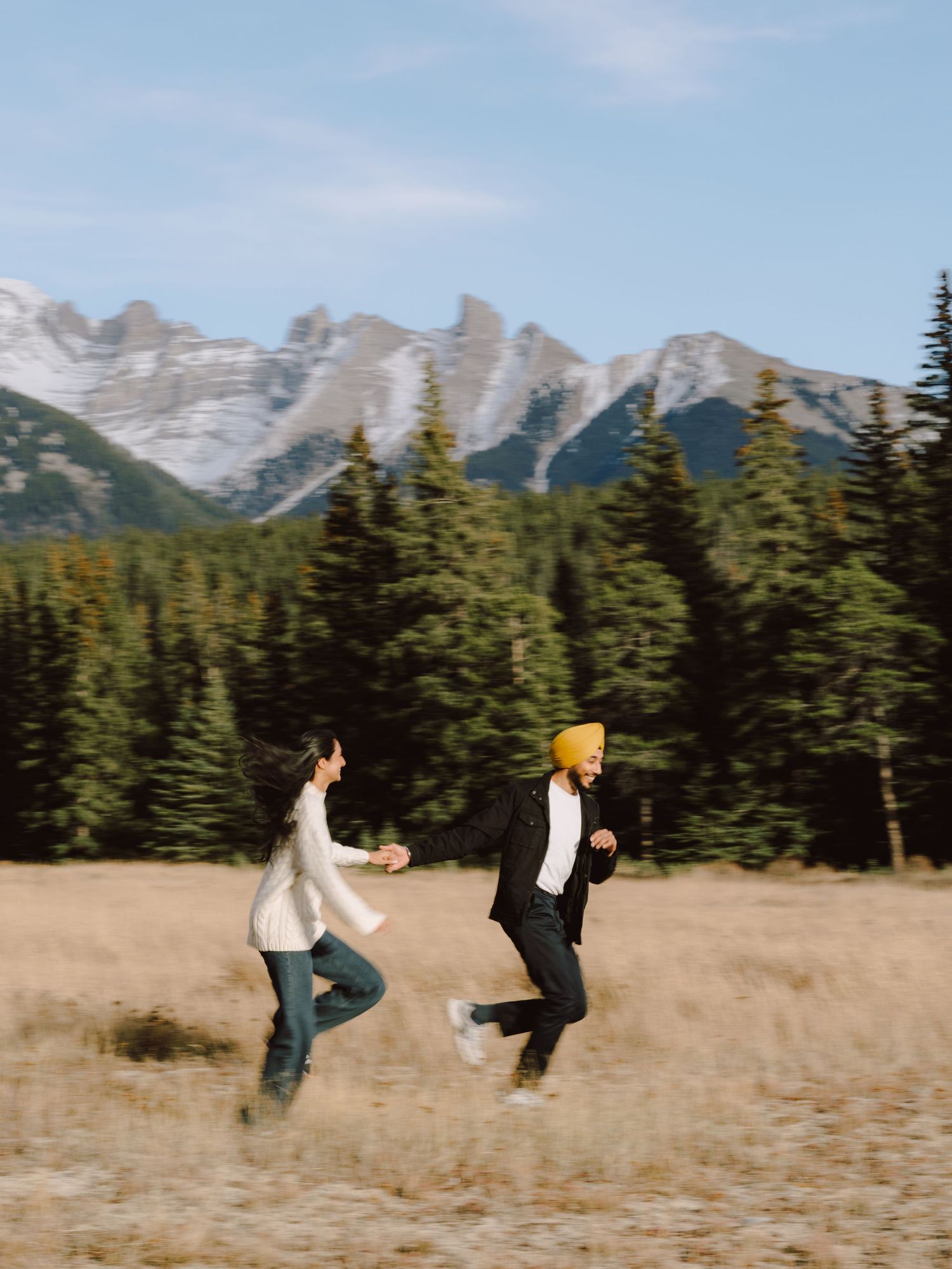 Joyful couple running through golden autumn meadow with snow-capped Rocky Mountains and pine trees behind.