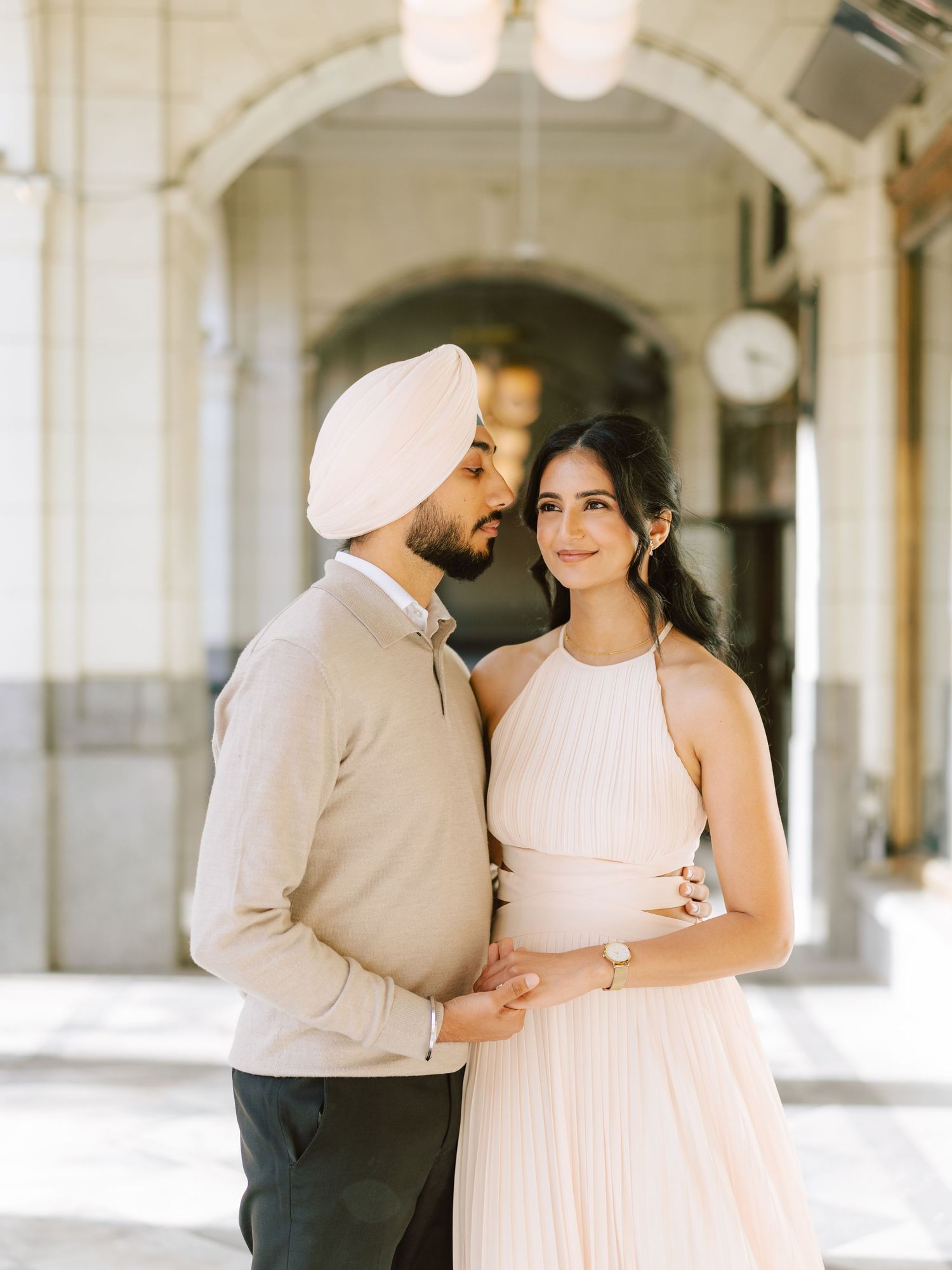 Sikh couple in elegant beige and blush outfits embrace under arched colonnade in sunlight.