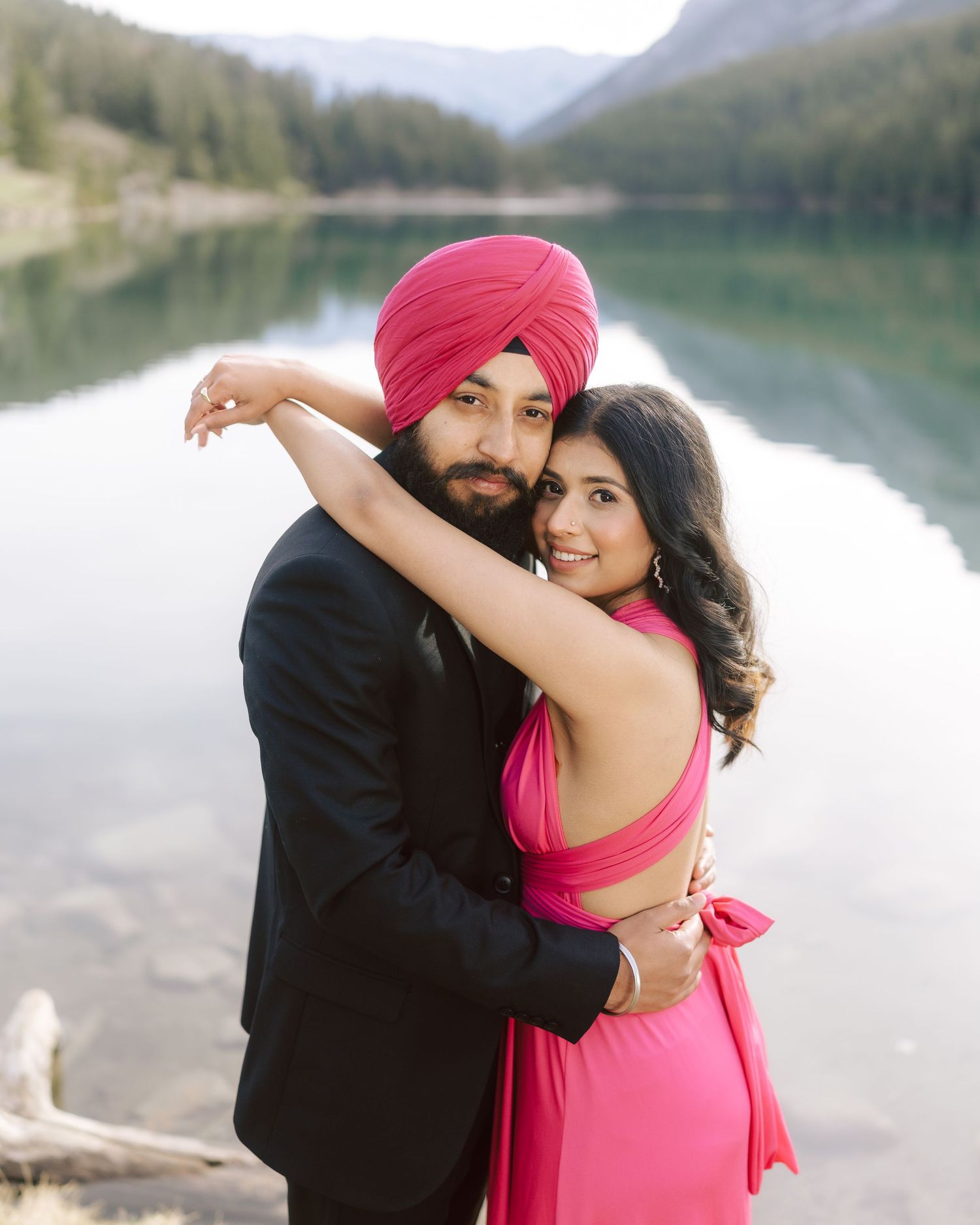 Couple embracing by reflective mountain lake, man in pink turban and black blazer, woman in pink dress.