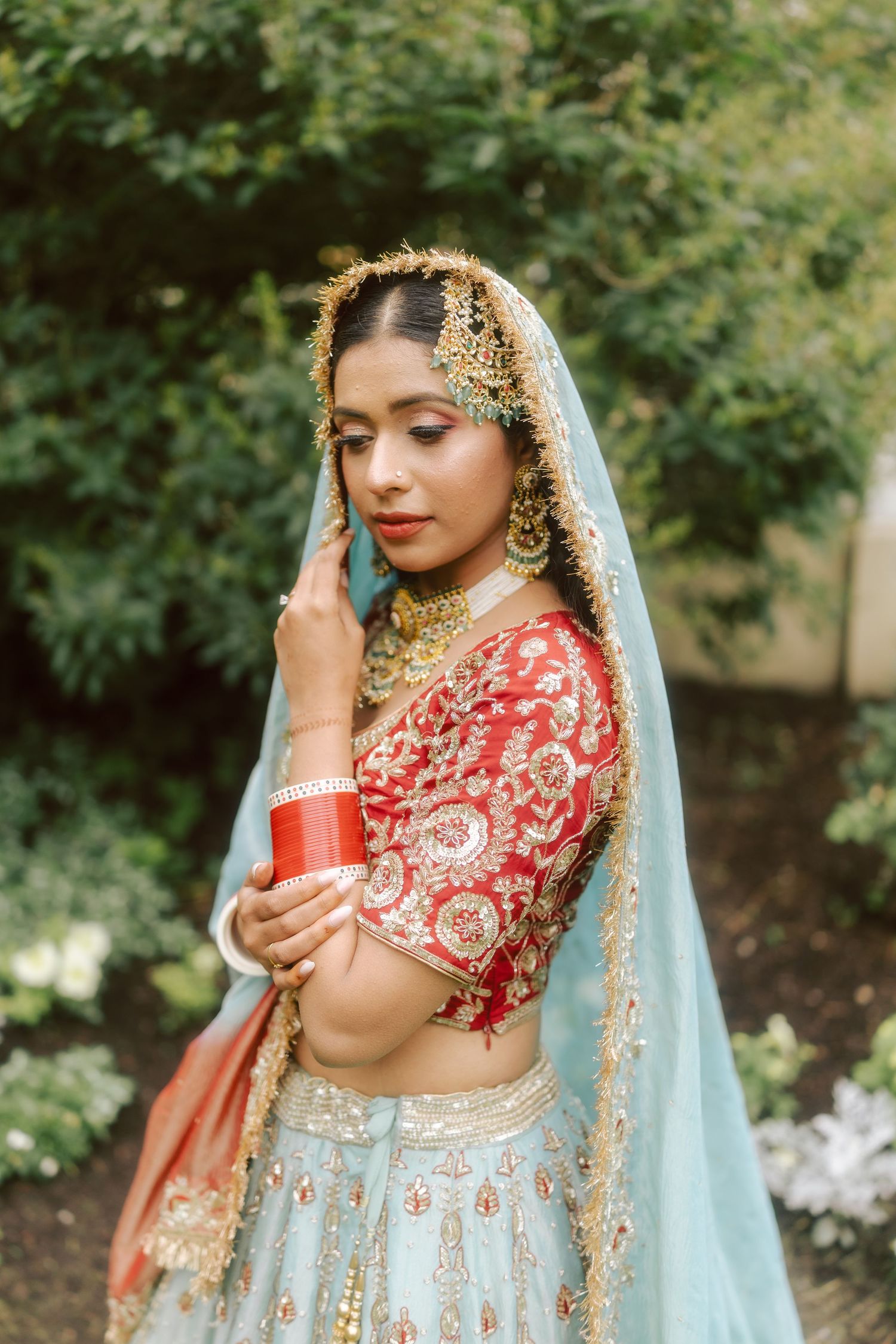 Indian bride in red and blue embroidered lehenga with gold maang tikka and dupatta in garden setting.