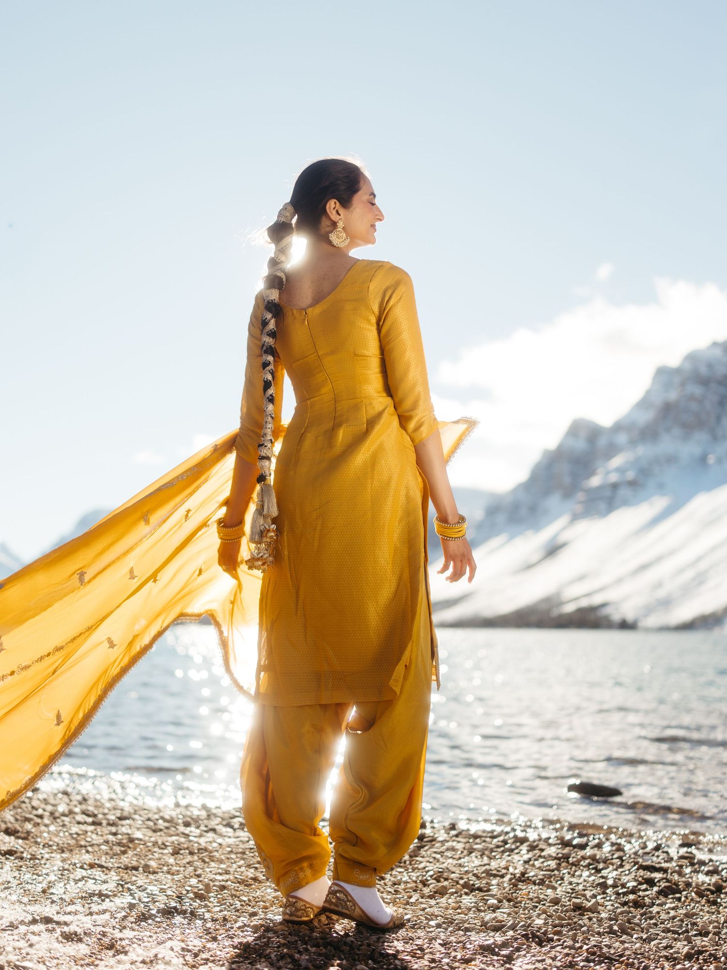 Woman in mustard yellow salwar kameez with flowing dupatta standing by snowy mountain lake.