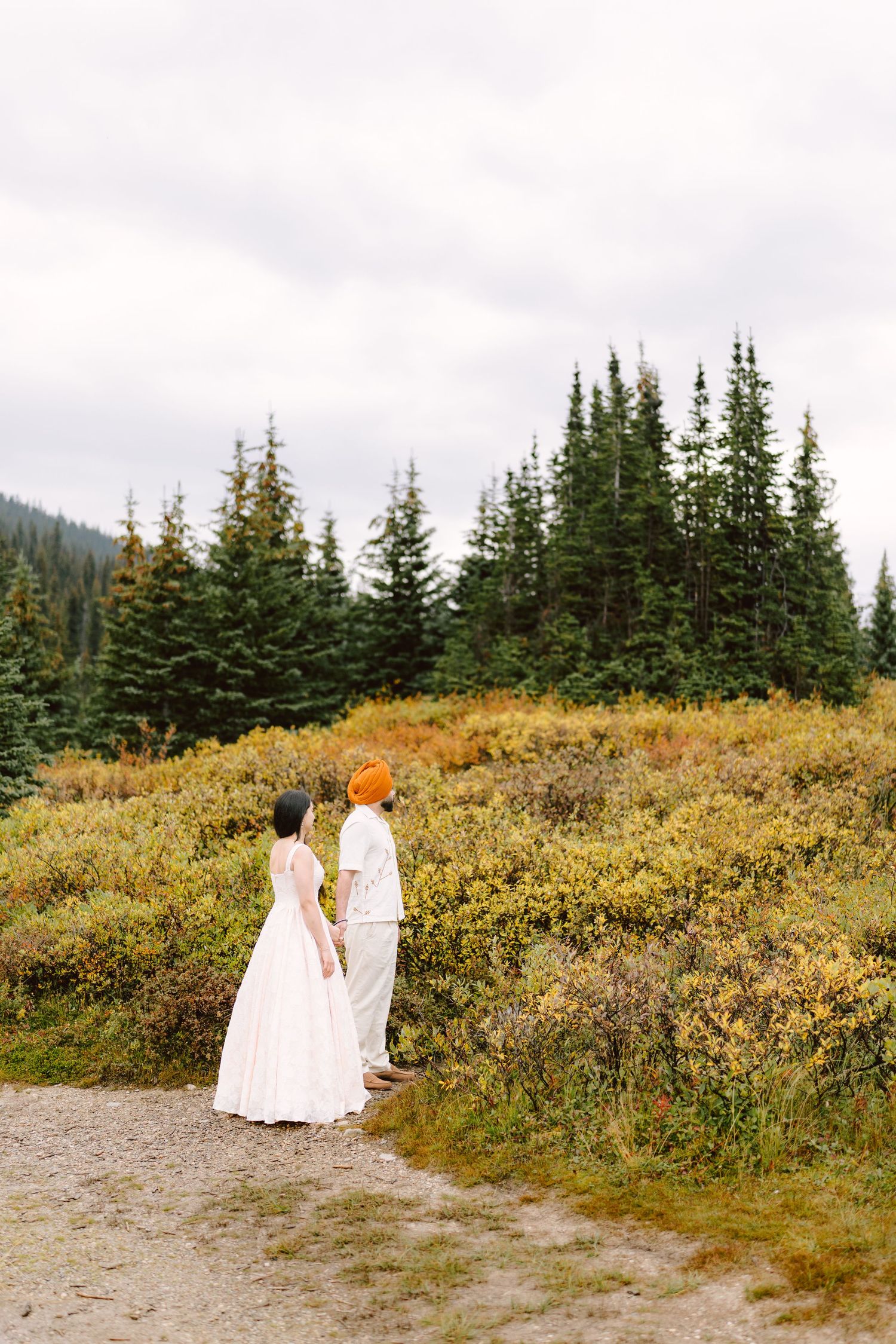 Couple holding hands on gravel path surrounded by autumn foliage and evergreen forest in the Rockies.