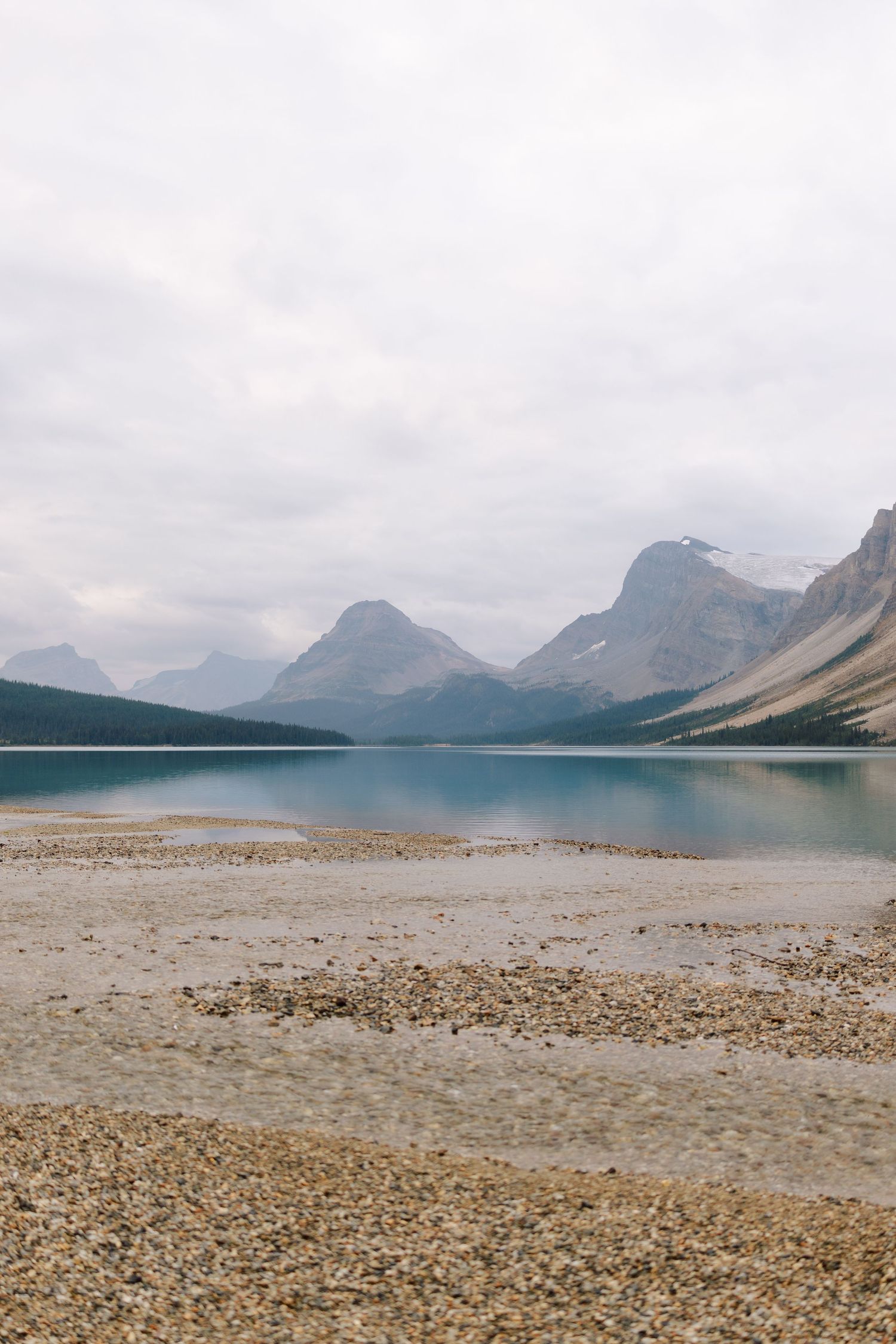 Peaceful Bow Lake with pebble shoreline, turquoise glacial water, and layered mountain peaks in Banff.