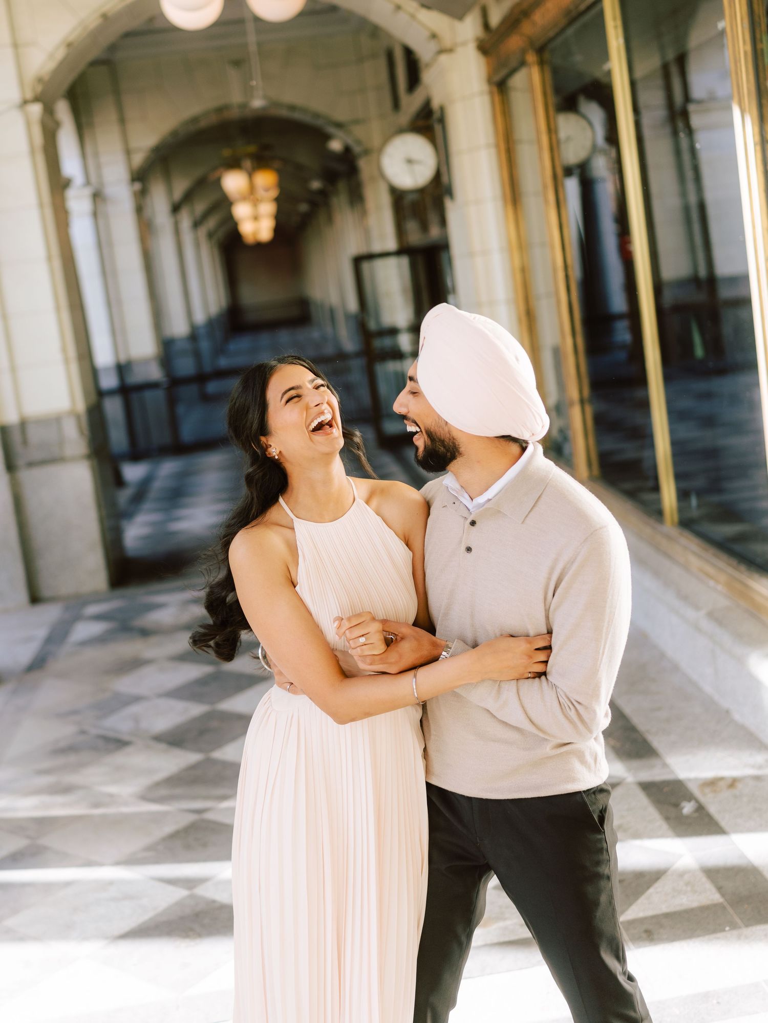 Laughing couple in elegant attire embrace in a sunlit architectural corridor during engagement photos.