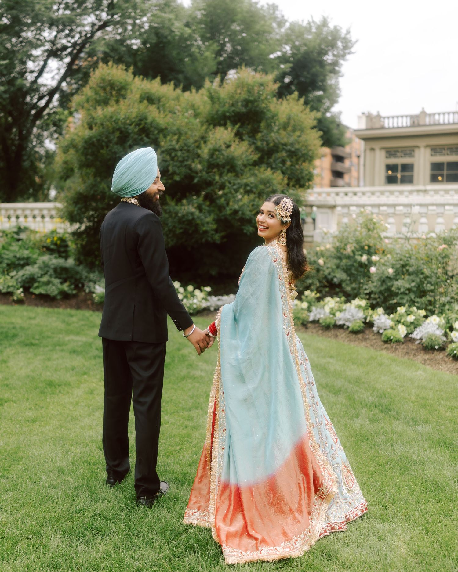Sikh couple holds hands in garden, bride in blue lehenga, groom in black suit.