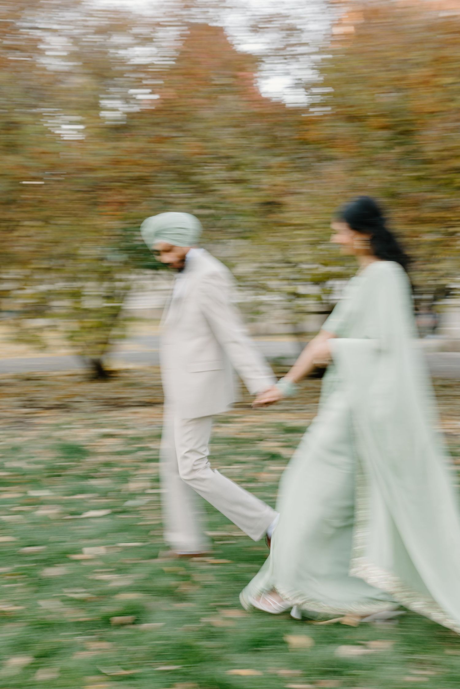 Blurred motion photo of a Sikh couple holding hands walking through an autumn park in matching sage outfits.