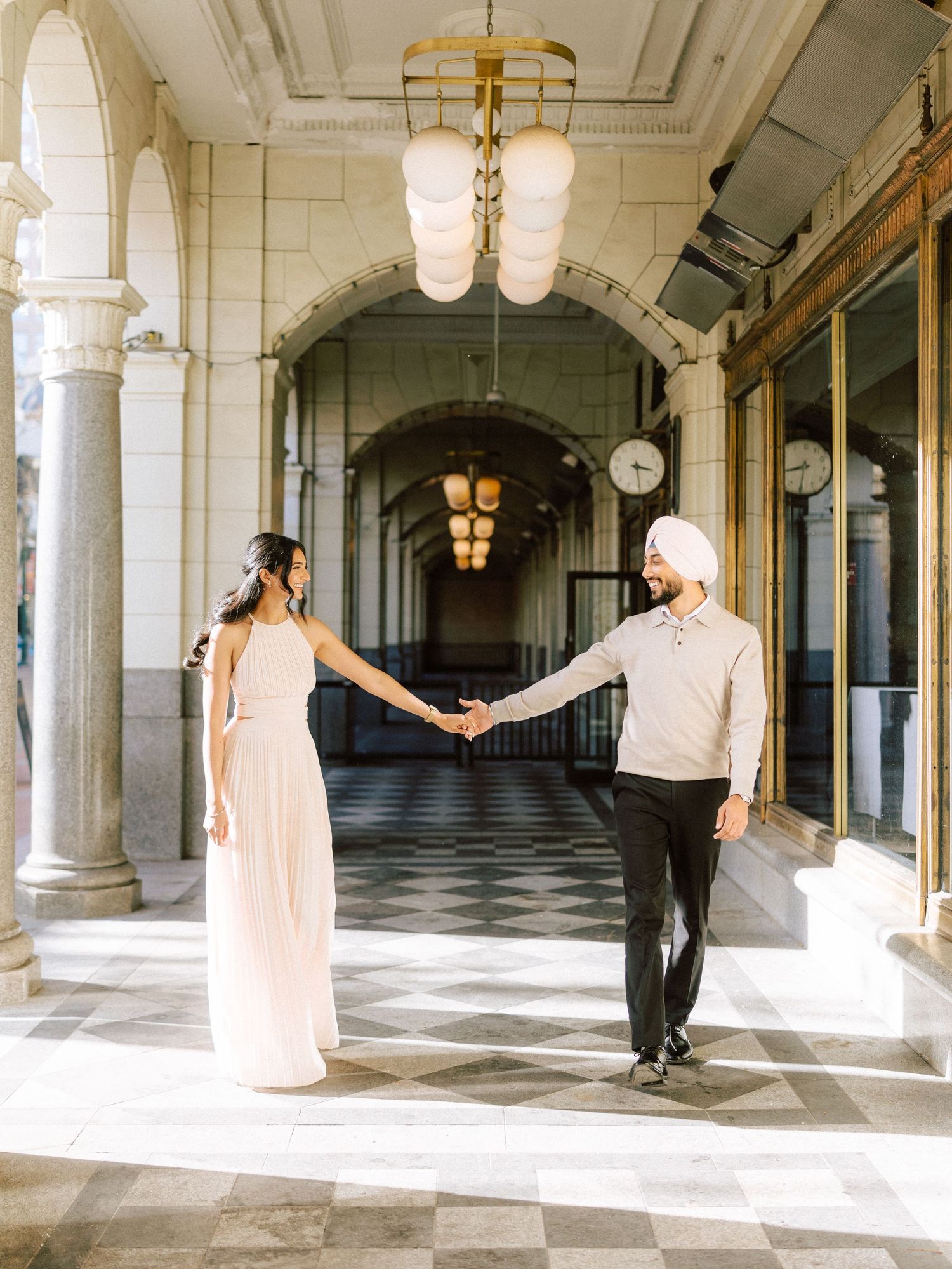 Sikh couple holding hands dancing in sunlit classical arcade with ornate chandeliers overhead.