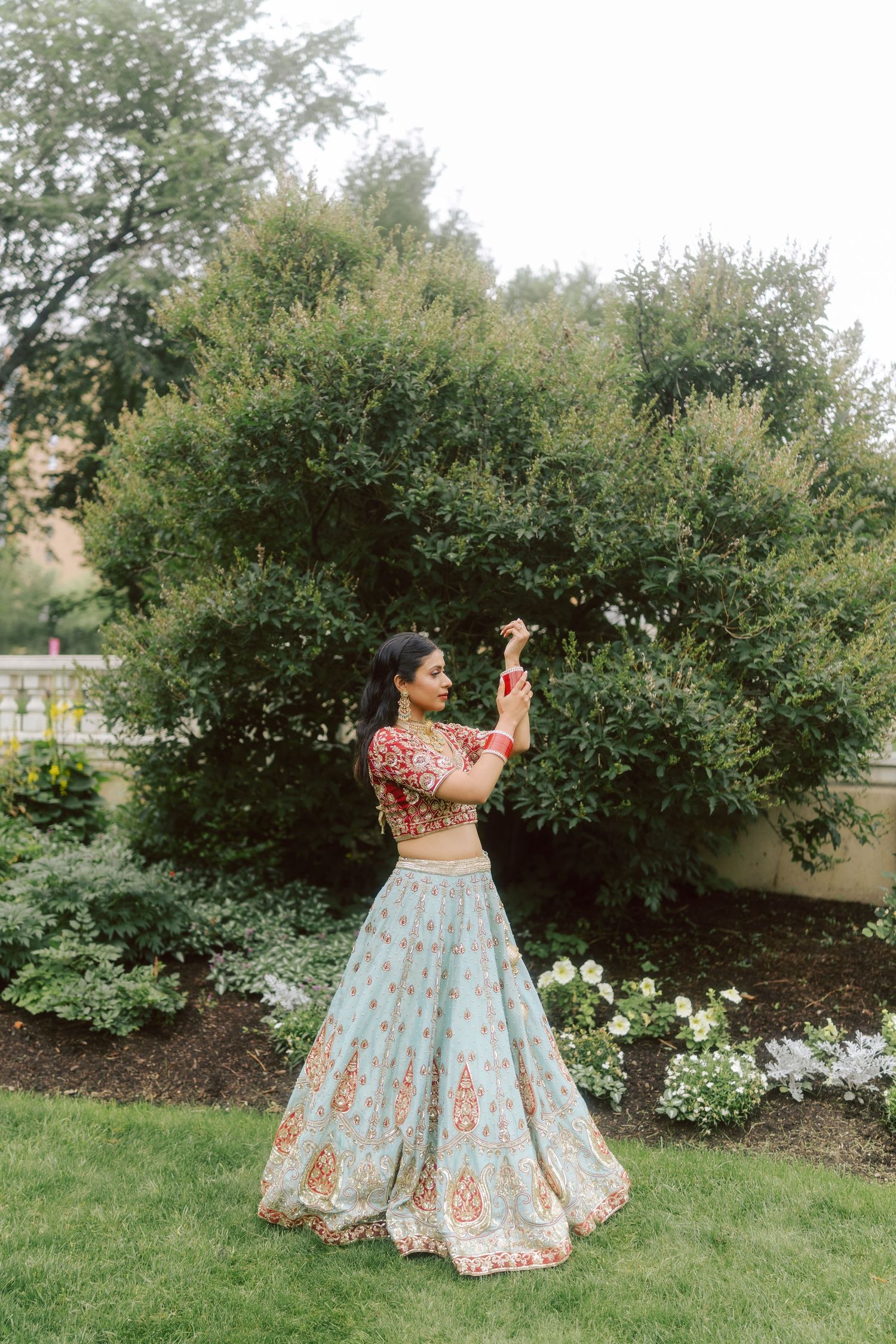 Bride in traditional red and blue lehenga twirls in lush garden setting.