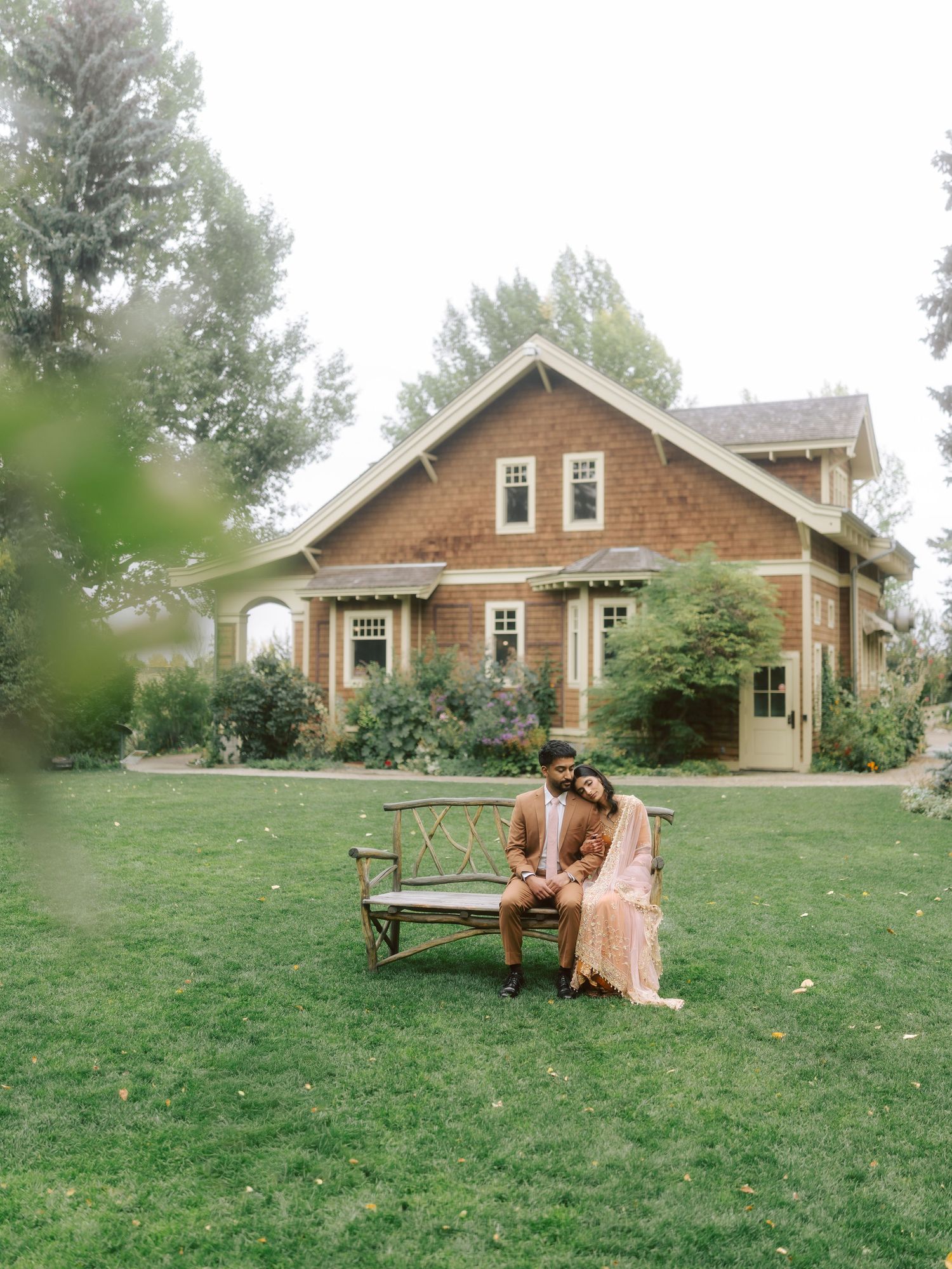 South Asian couple sitting on wooden bench in front of charming craftsman house surrounded by greenery.