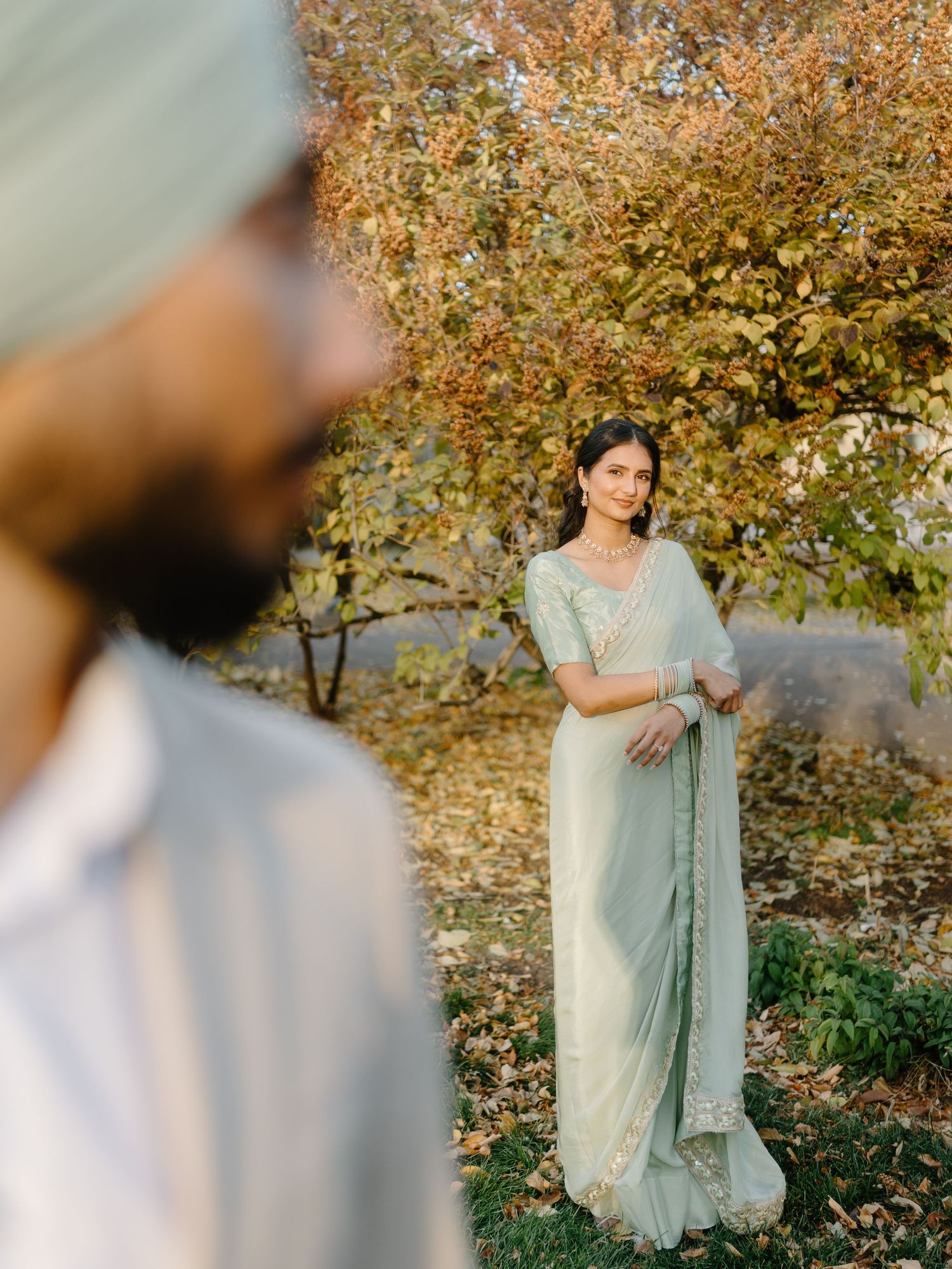 Beautiful bride in mint green saree smiles softly at groom in autumn garden setting.