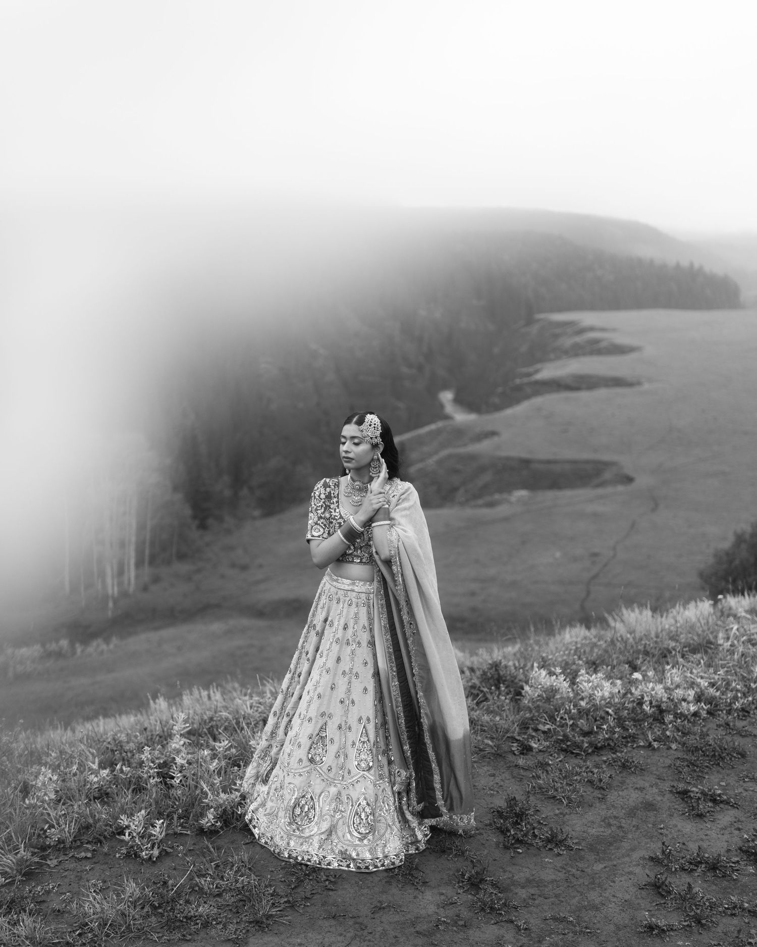 Black and white portrait of Sikh bride in embroidered lehenga standing on foggy mountain overlook.