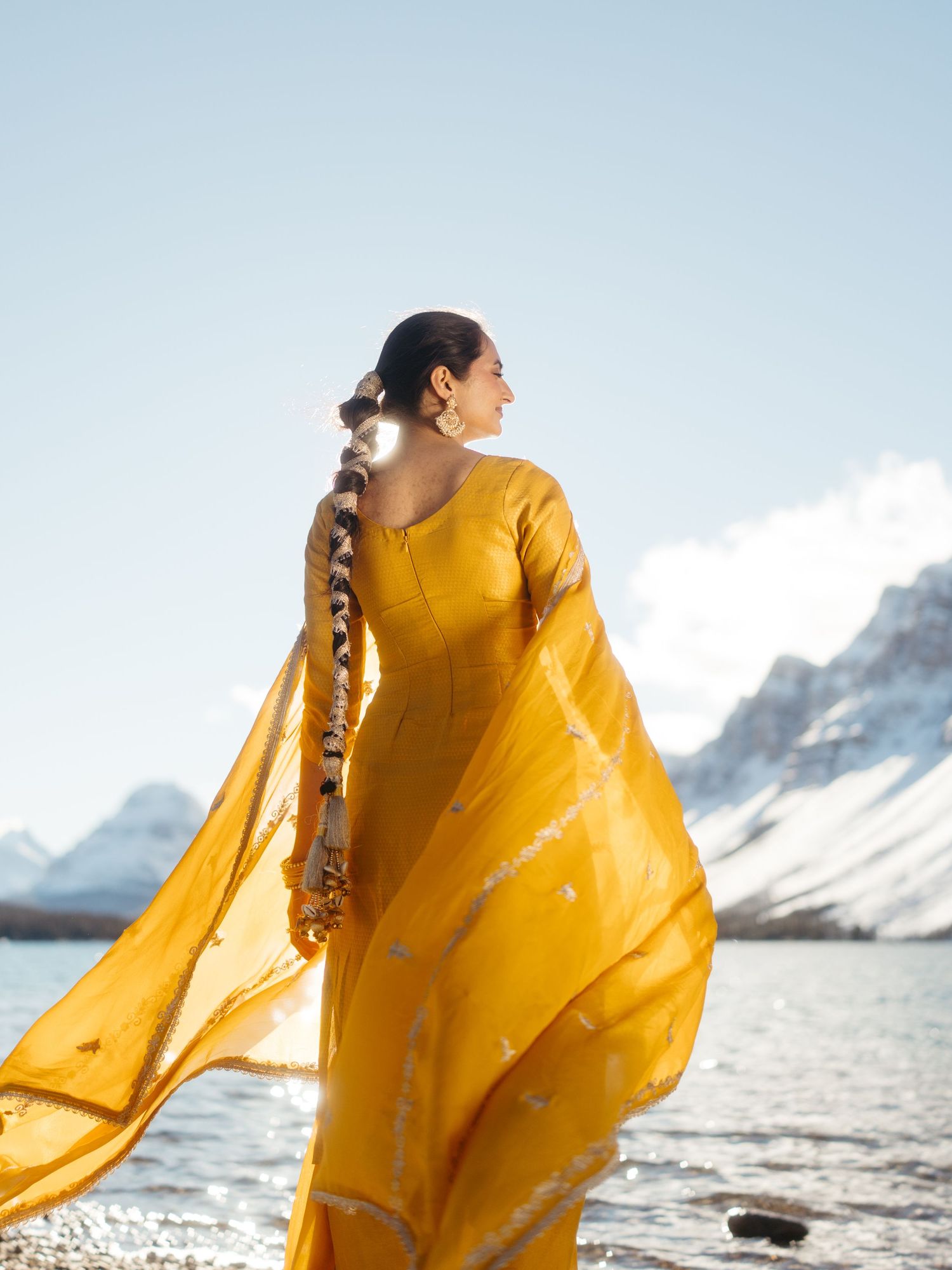 Woman in vibrant yellow salwar suit with braided hair adorned with flowers by mountain lake.
