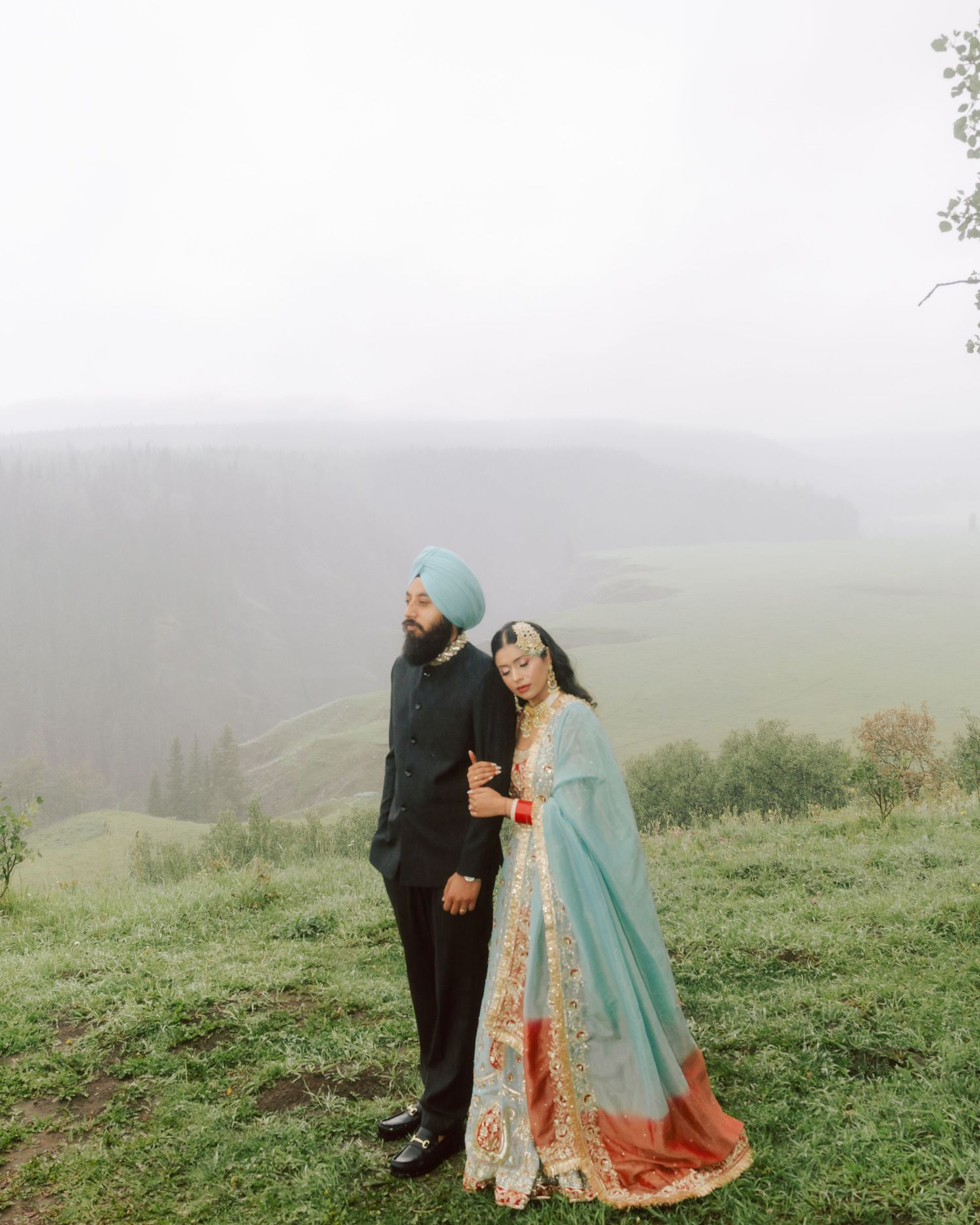 Sikh bride and groom in traditional blue and red lehenga holding hands on misty mountain hilltop.