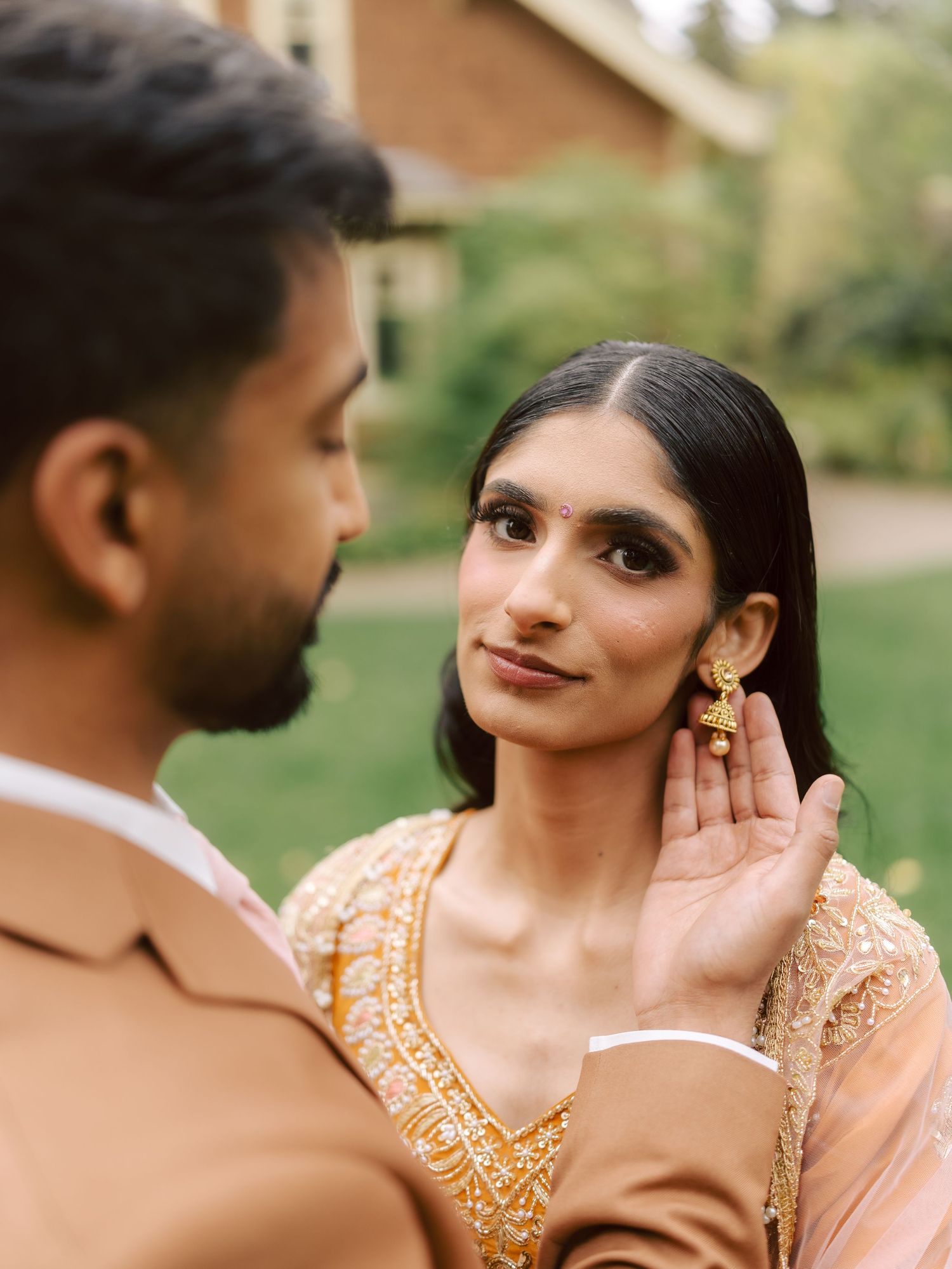South Asian groom tenderly touching bride's face as she smiles, wearing gold jhumka earrings.