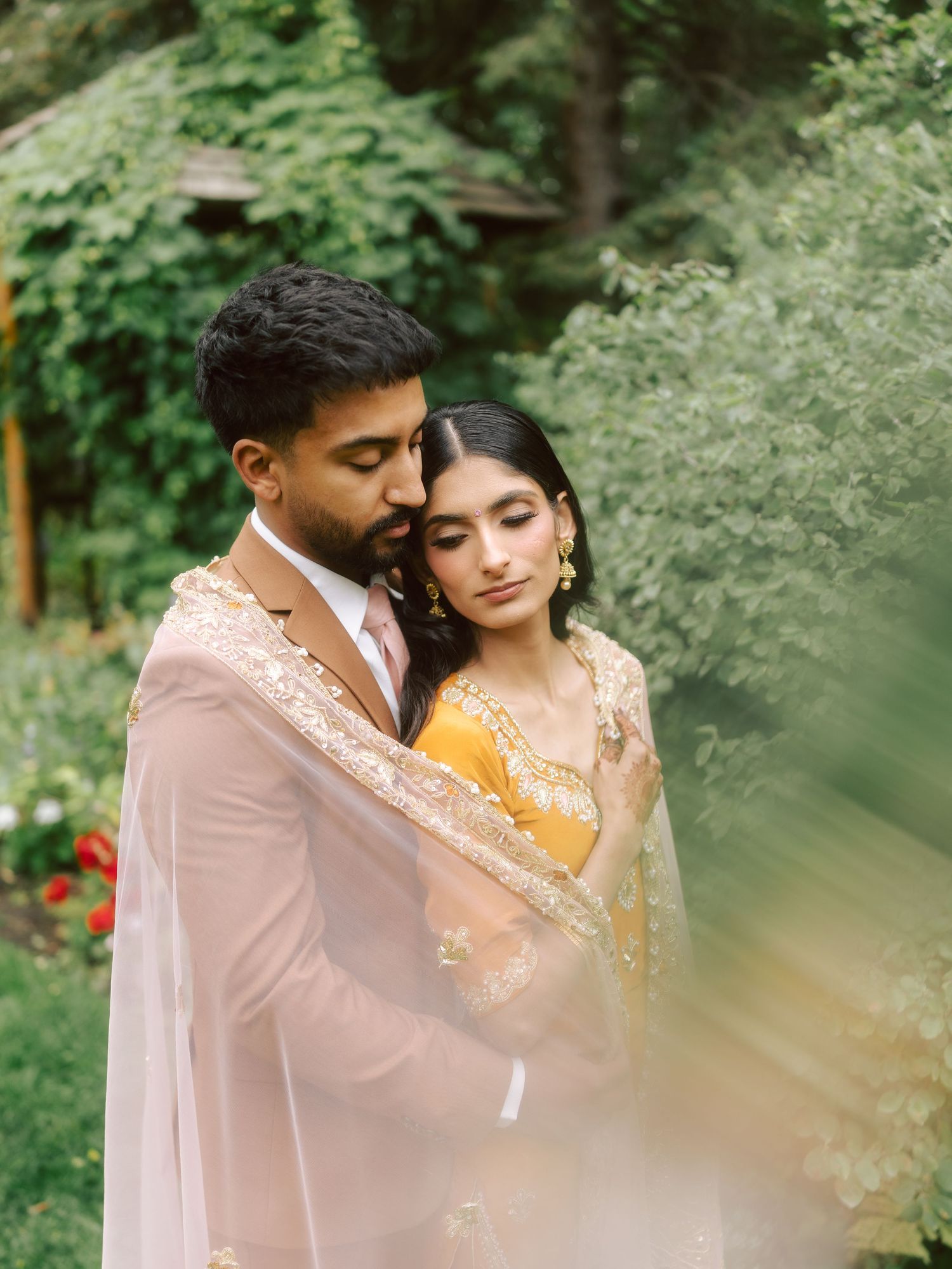 South Asian bride and groom in traditional attire share intimate embrace in lush garden setting.