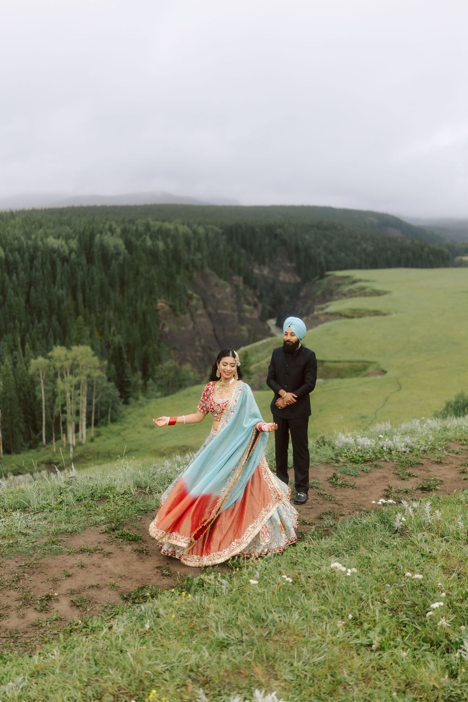 Indian bride in blue and red lehenga twirling while groom in black watches against scenic valley backdrop.