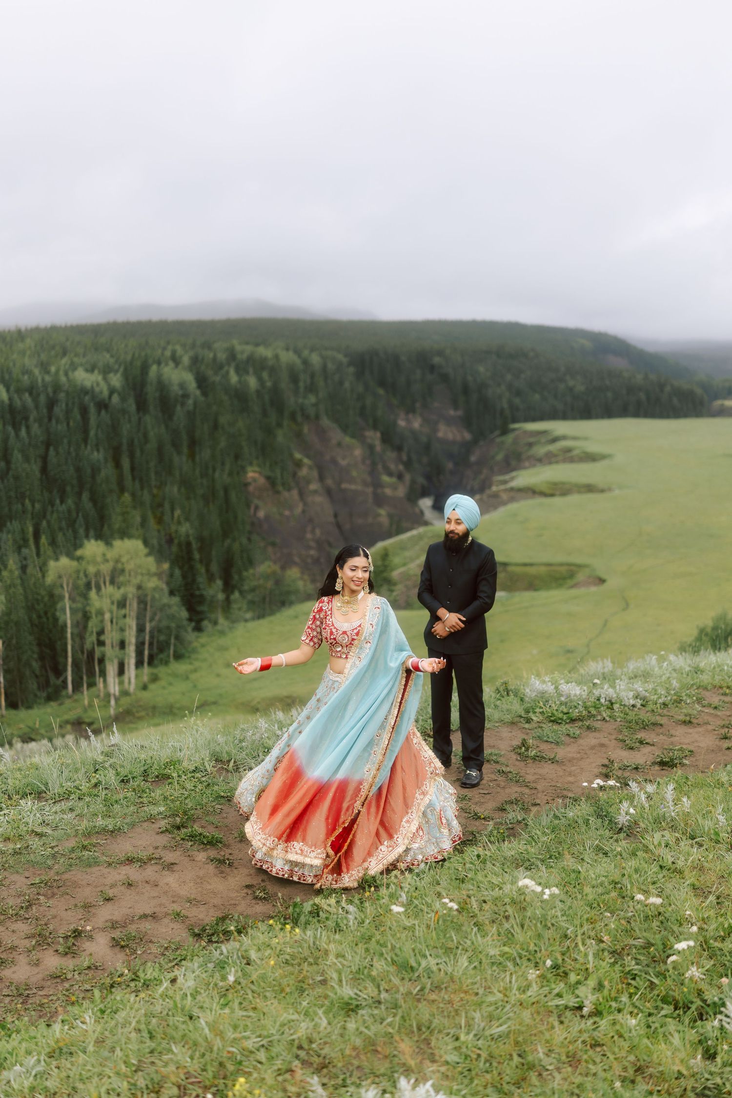 Sikh bride spinning in ornate blue red lehenga as turbaned groom watches on lush hilltop overlook.