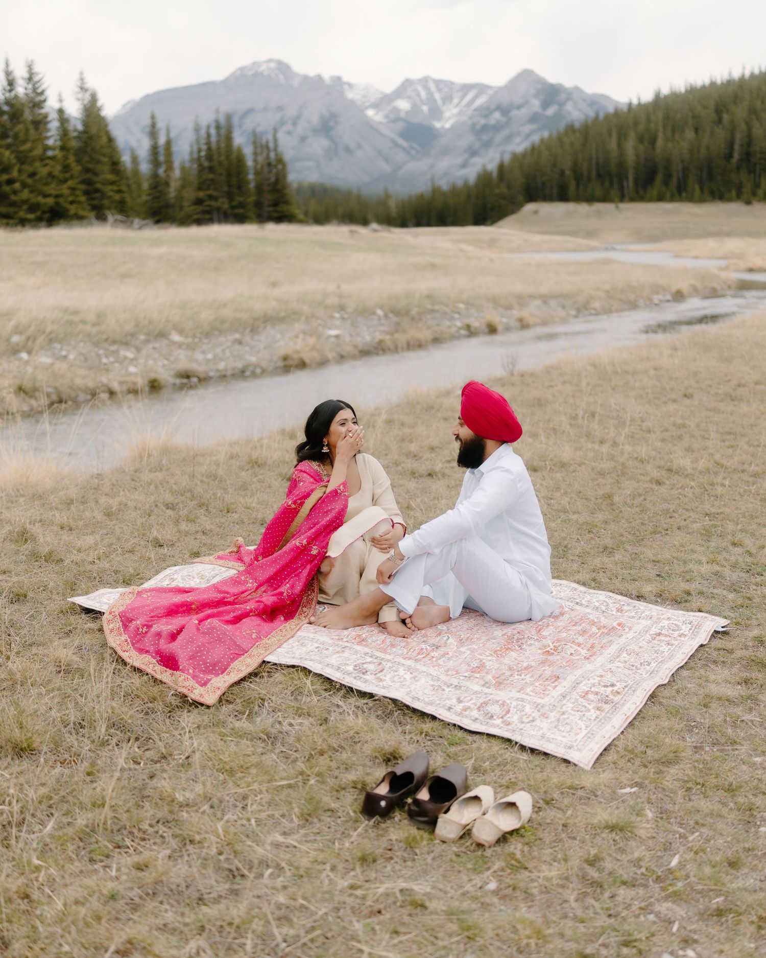 A Sikh couple sits on a vintage rug in a mountain meadow, woman in pink lehenga and man in white sherwani with red turban.