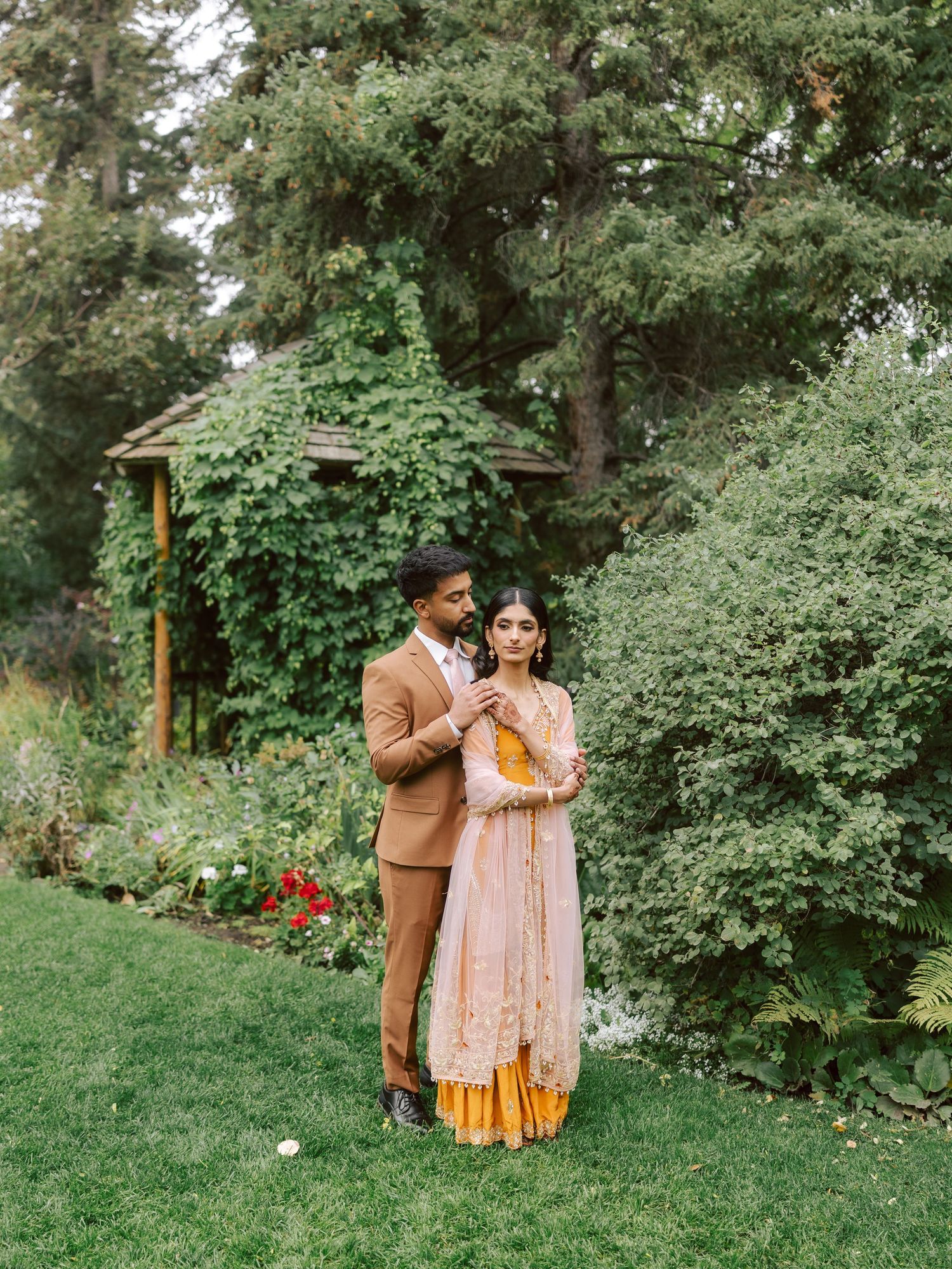 South Asian groom in tan suit stands with bride in pink lehenga in lush garden setting.