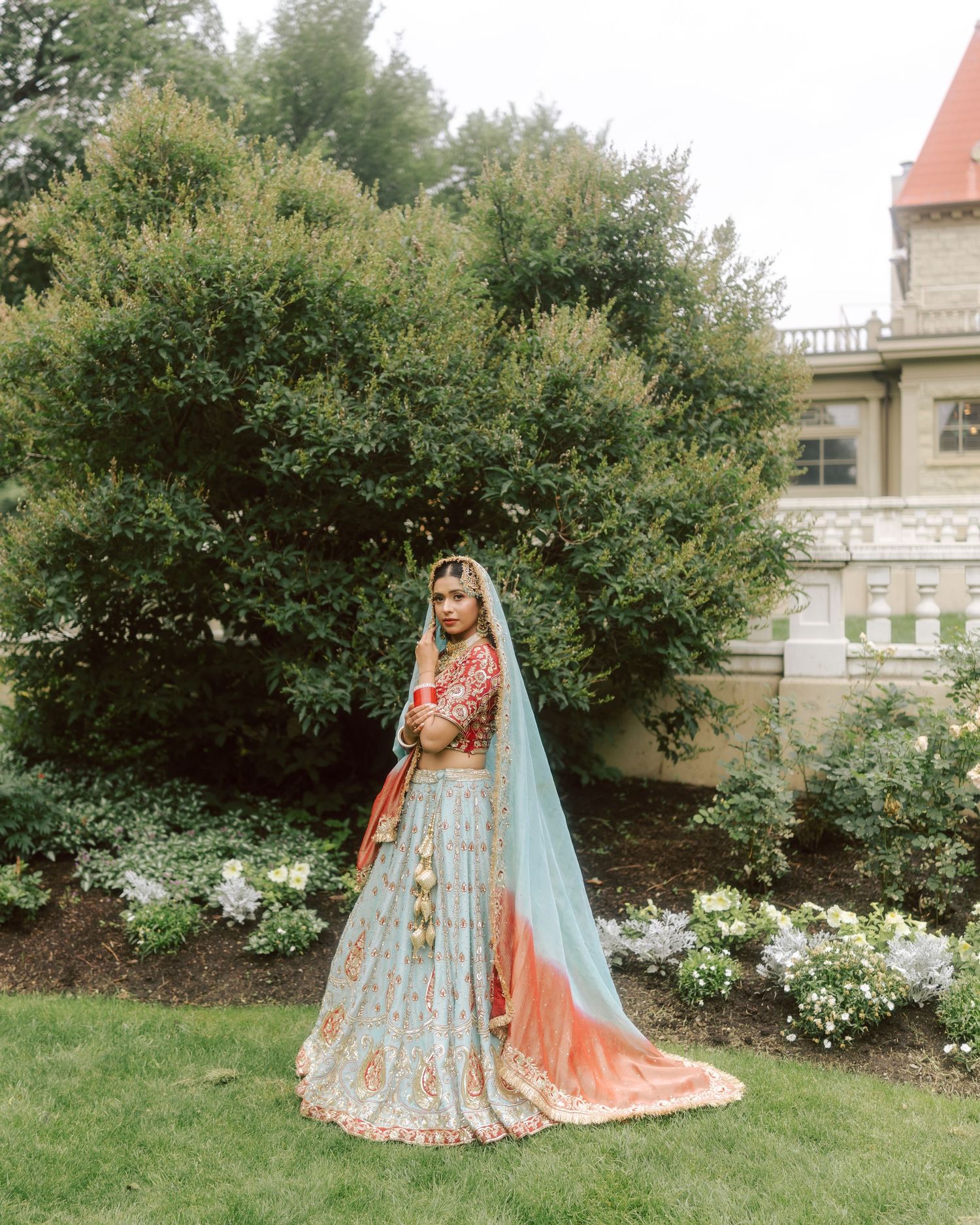 Indian bride in blue and red lehenga stands before historic castle garden.