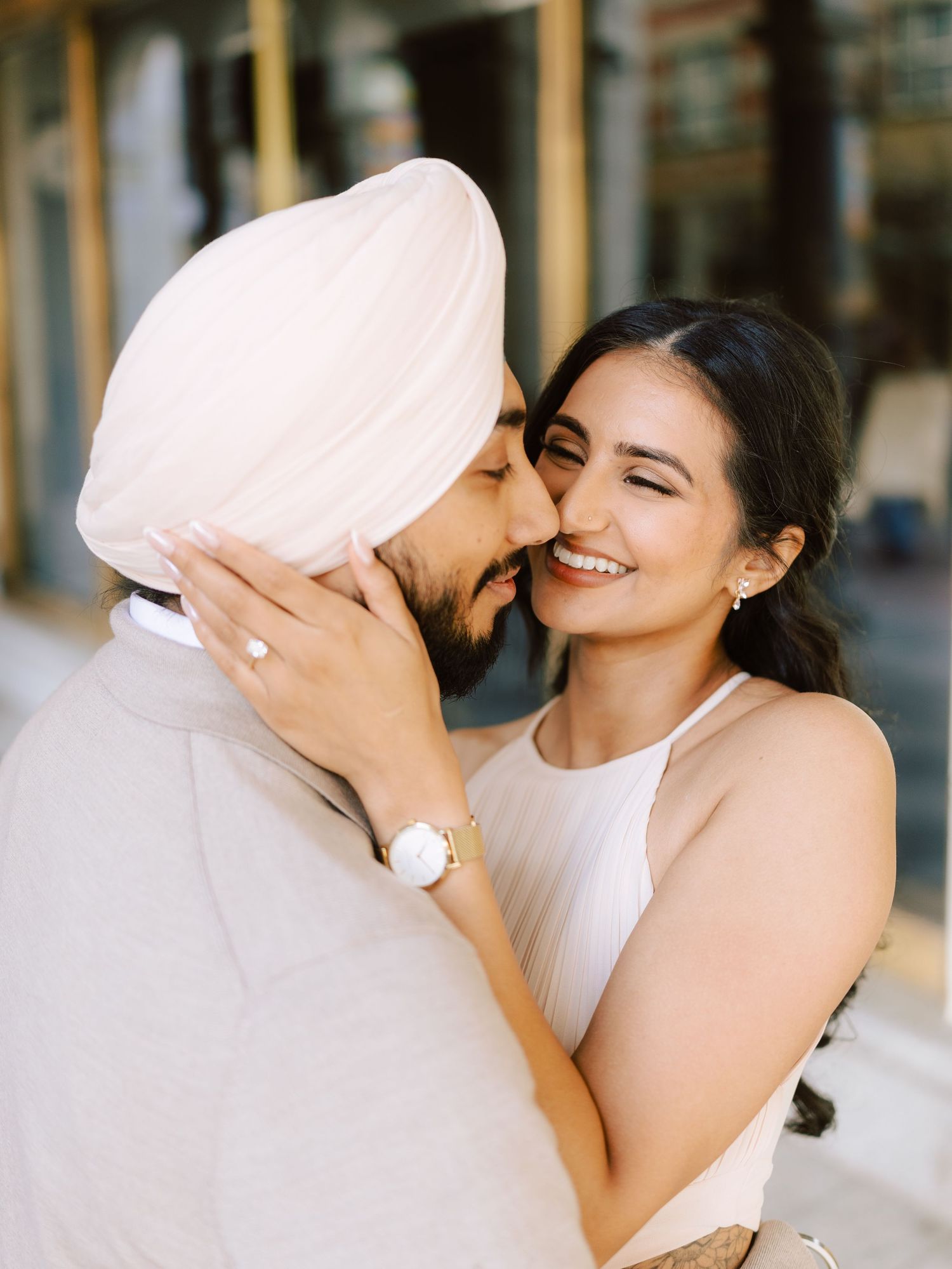 Groom in pink turban kisses smiling bride on the cheek during outdoor engagement session.