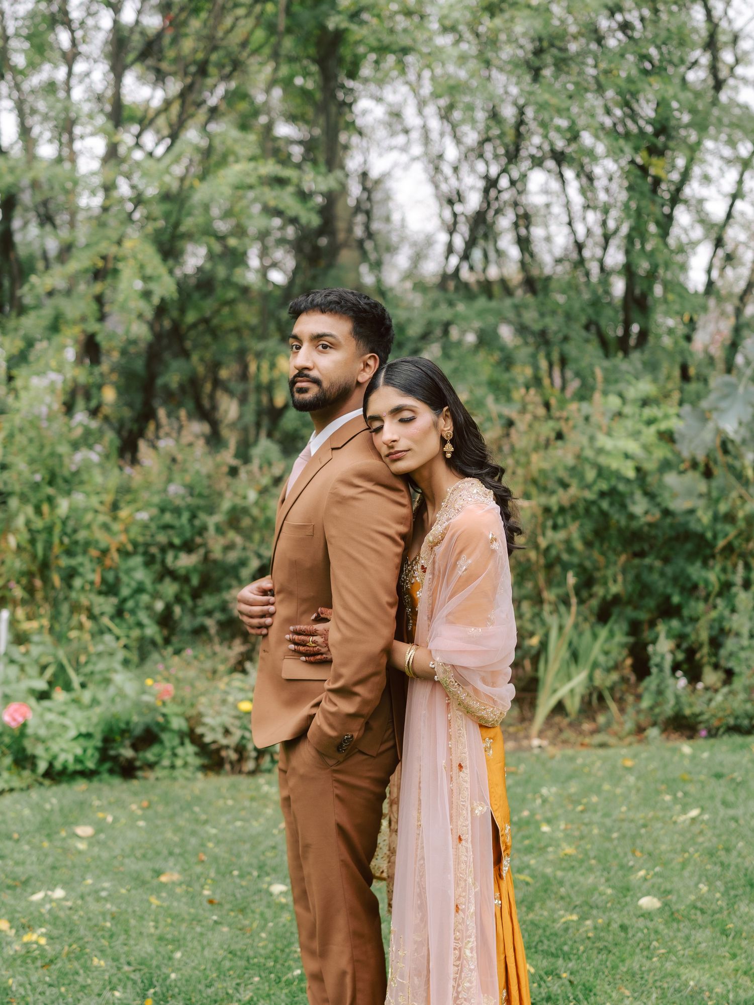 South Asian couple in traditional wedding attire embrace back-to-back in green outdoor garden.