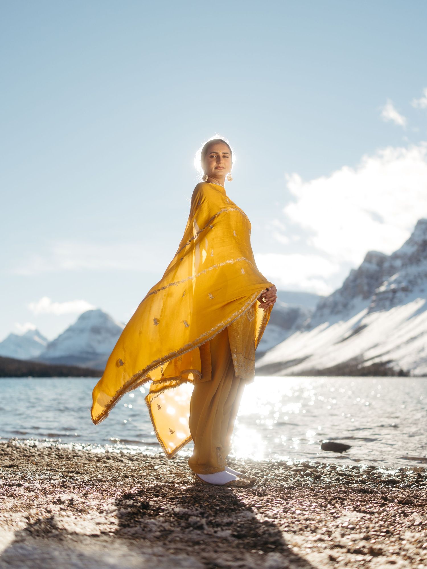 Woman in yellow salwar kameez with flowing dupatta posing by scenic snowy mountain lake.