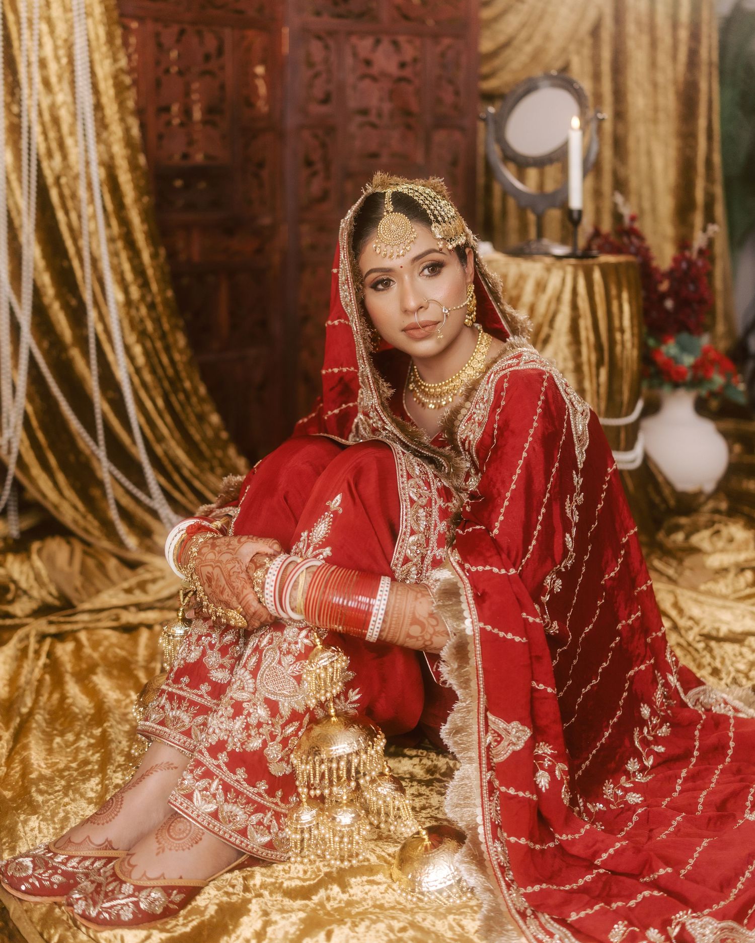Indian bride in red and gold embroidered lehenga with traditional jewelry, seated on golden fabric backdrop.
