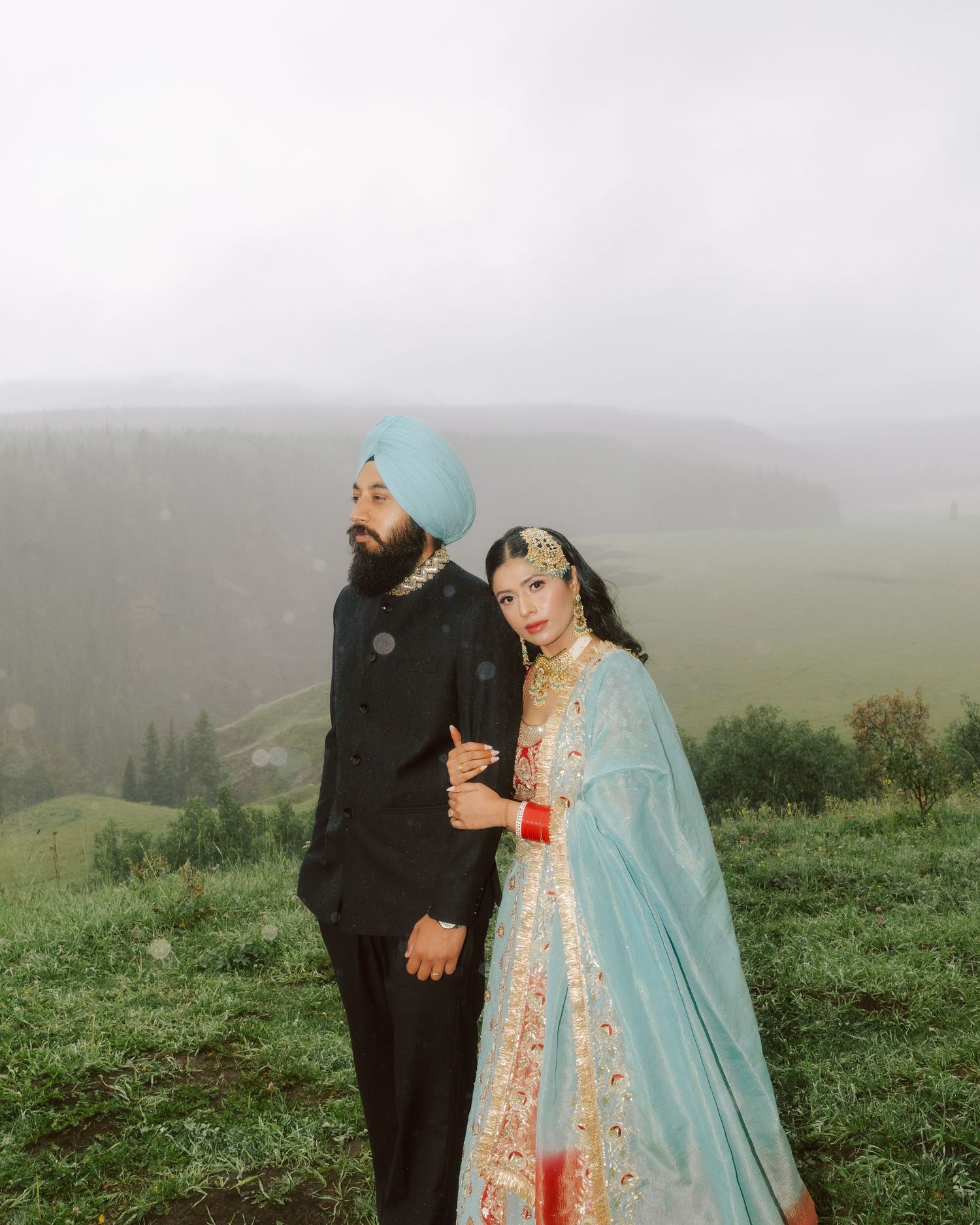 Sikh bride in blue lehenga and groom in black sherwani posing on misty mountain overlook.