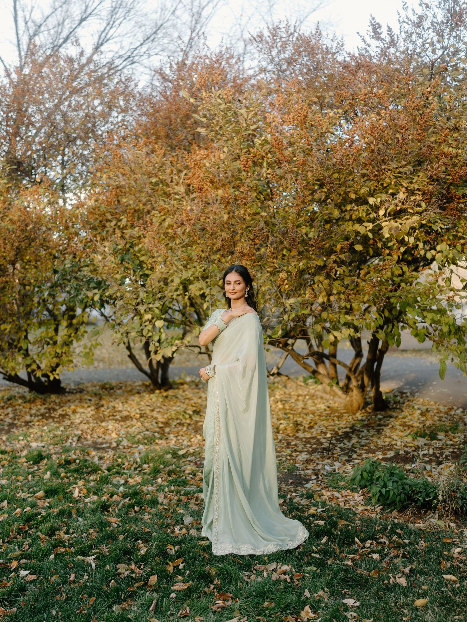 Elegant bride in mint green saree poses gracefully among colorful autumn trees in outdoor setting.