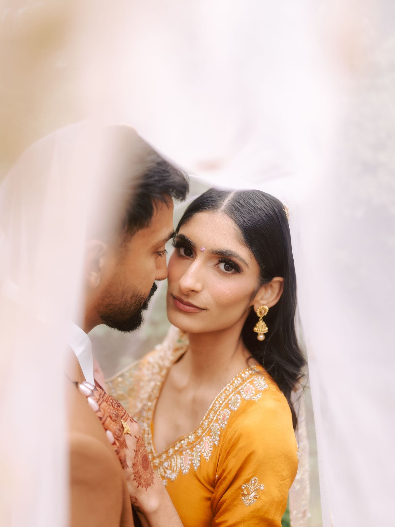Intimate portrait of South Asian bride in yellow lehenga with gold jewelry beneath a flowing veil.