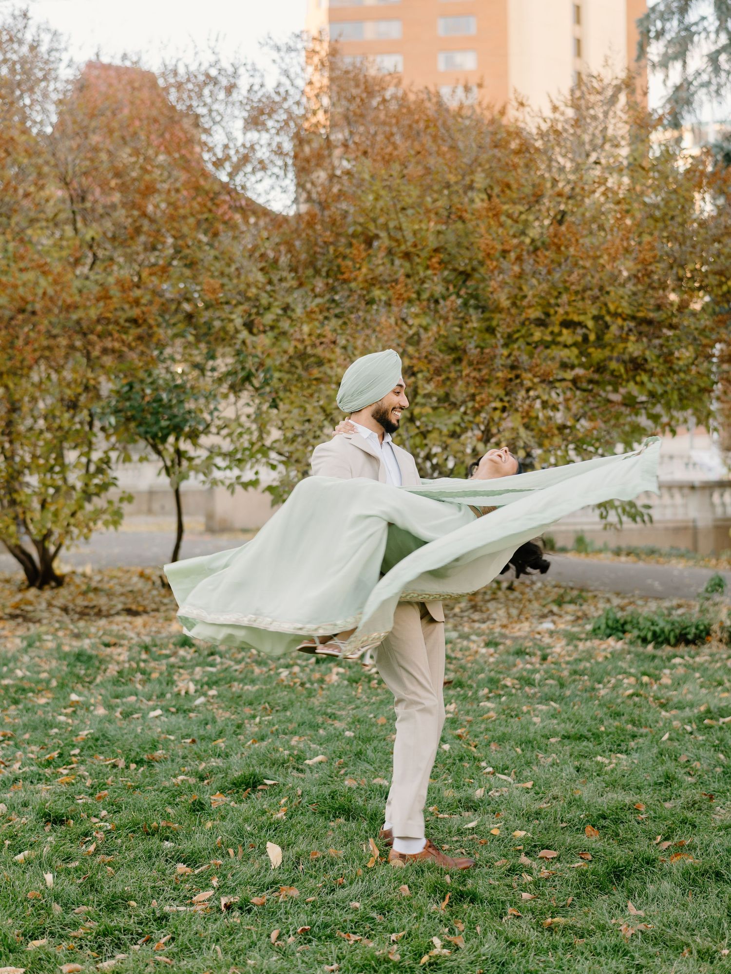 Joyful Sikh groom lifts bride in flowing mint saree in a playful autumn park moment.