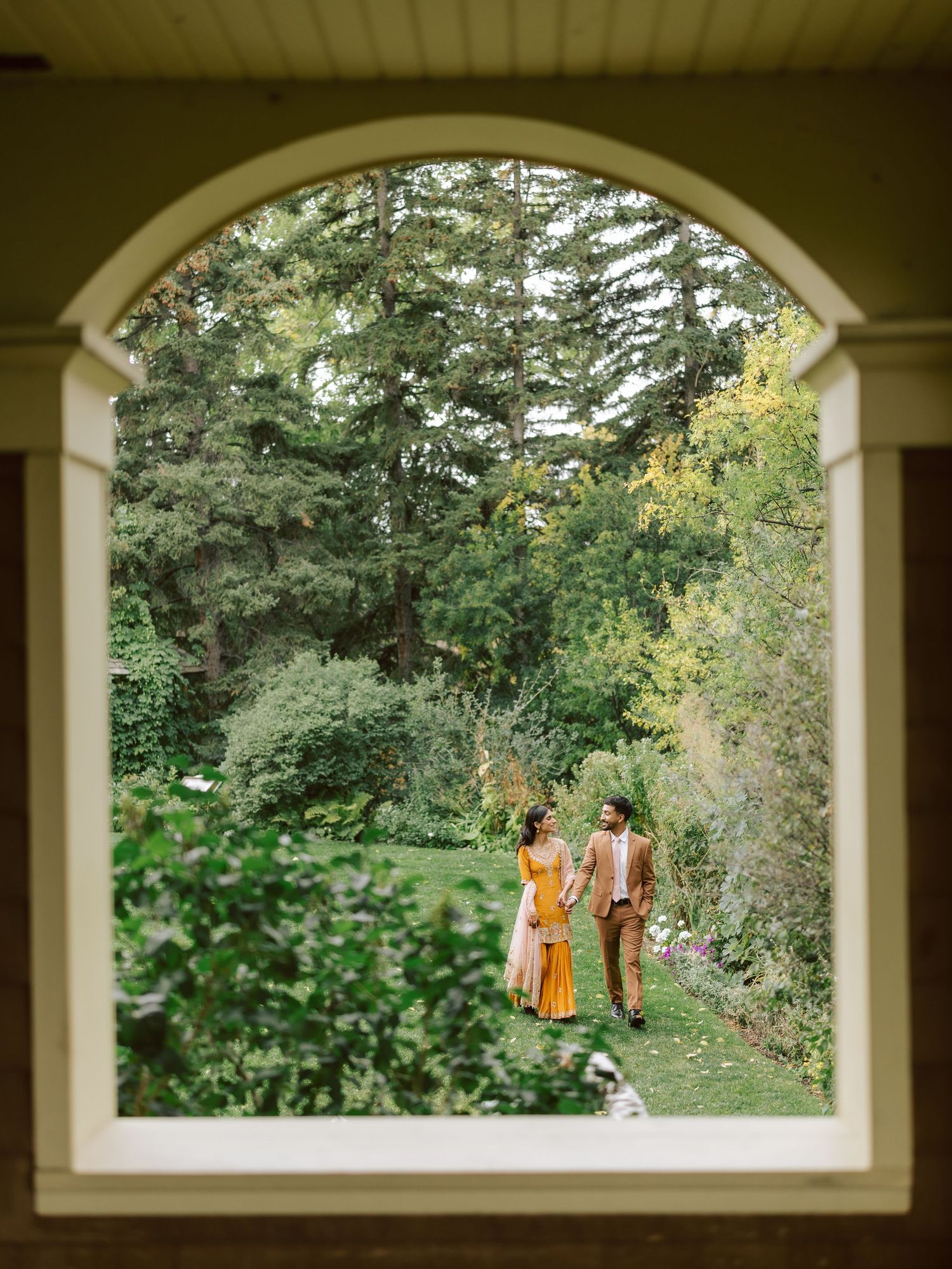 South Asian couple walking in garden viewed through arched window frame, bride in yellow lehenga.