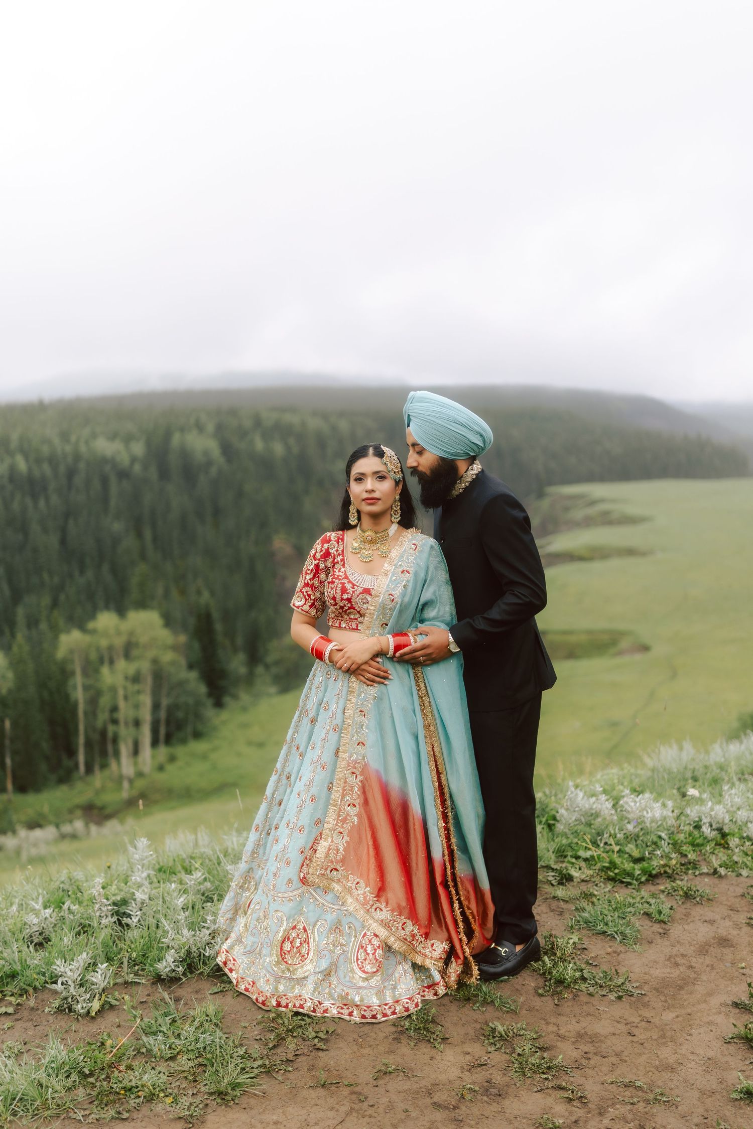 Sikh groom in black sherwani kissing bride in blue lehenga on scenic misty mountain overlook.