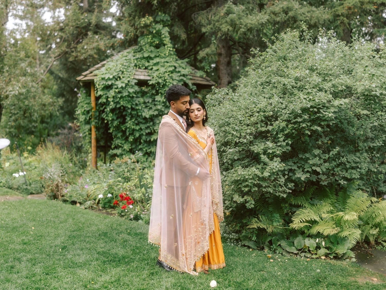 A couple in traditional Indian outfits embrace in a garden with a wooden arbor and colorful flowers.